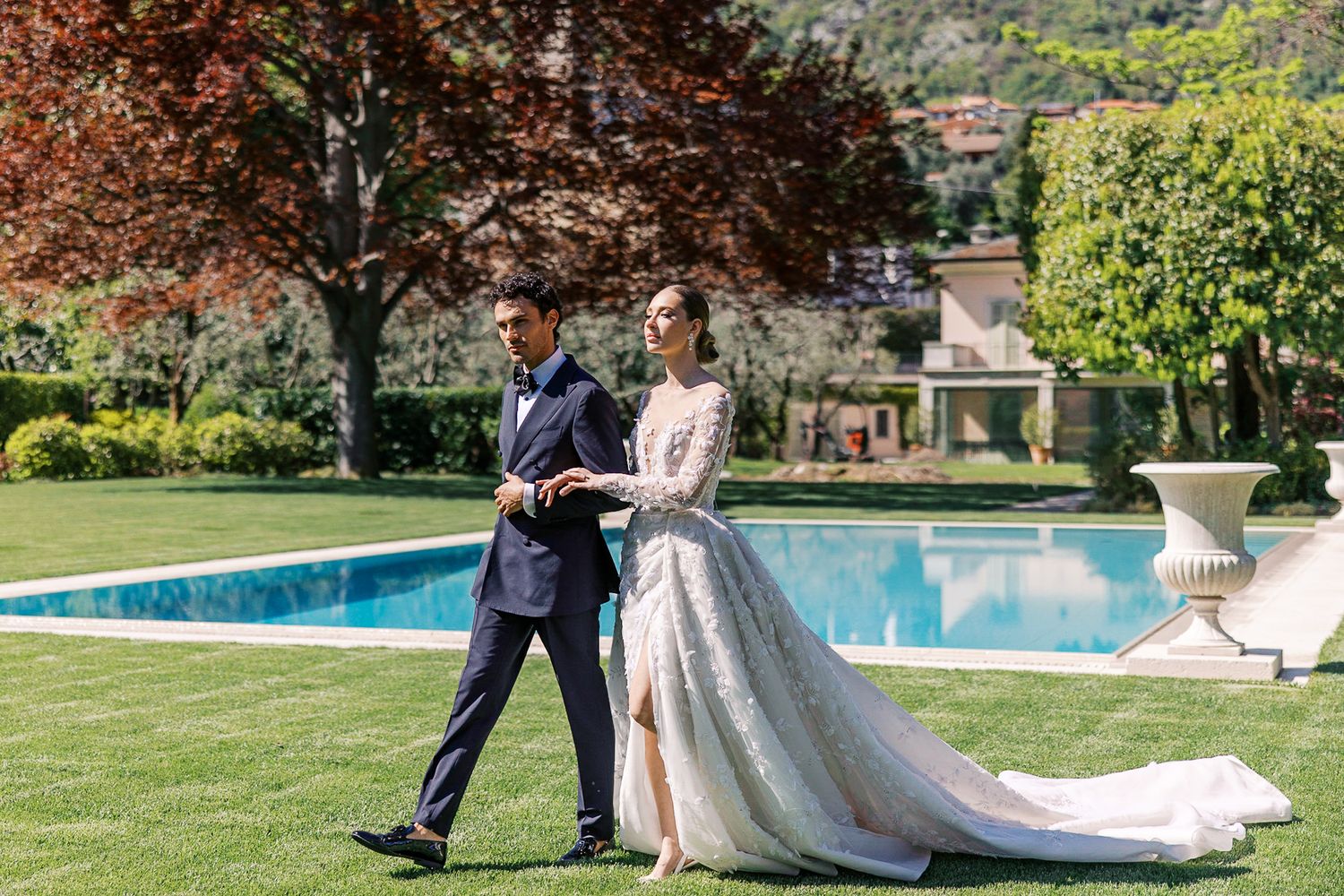 Elegant couple poses in formal attire by a swimming pool in a lush garden setting with ornate stone features at Lake Como Villa Balbiano.