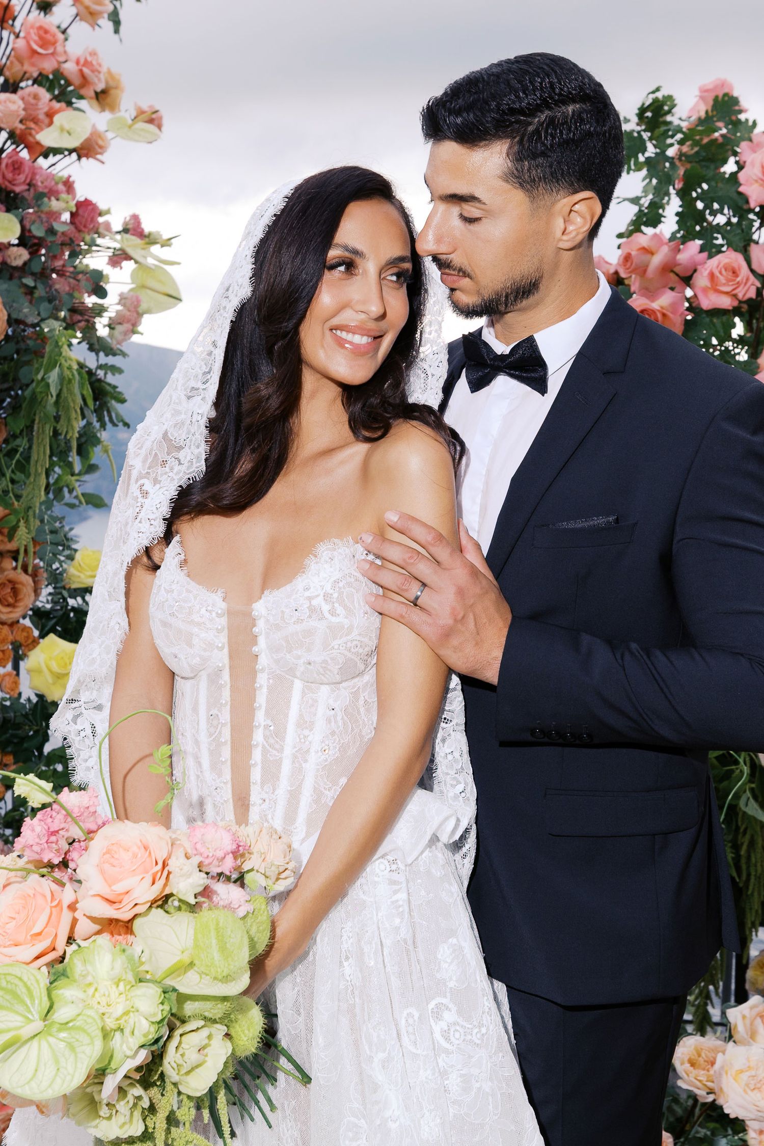 A bride in a white lace gown and groom in black tuxedo pose together under floral arch with peach and green blooms on Amalfi Coast.