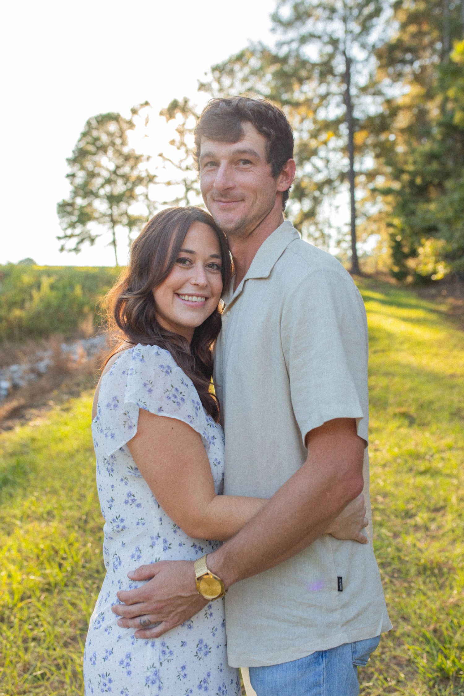 Couple embracing on grassy hillside with pine trees in background.