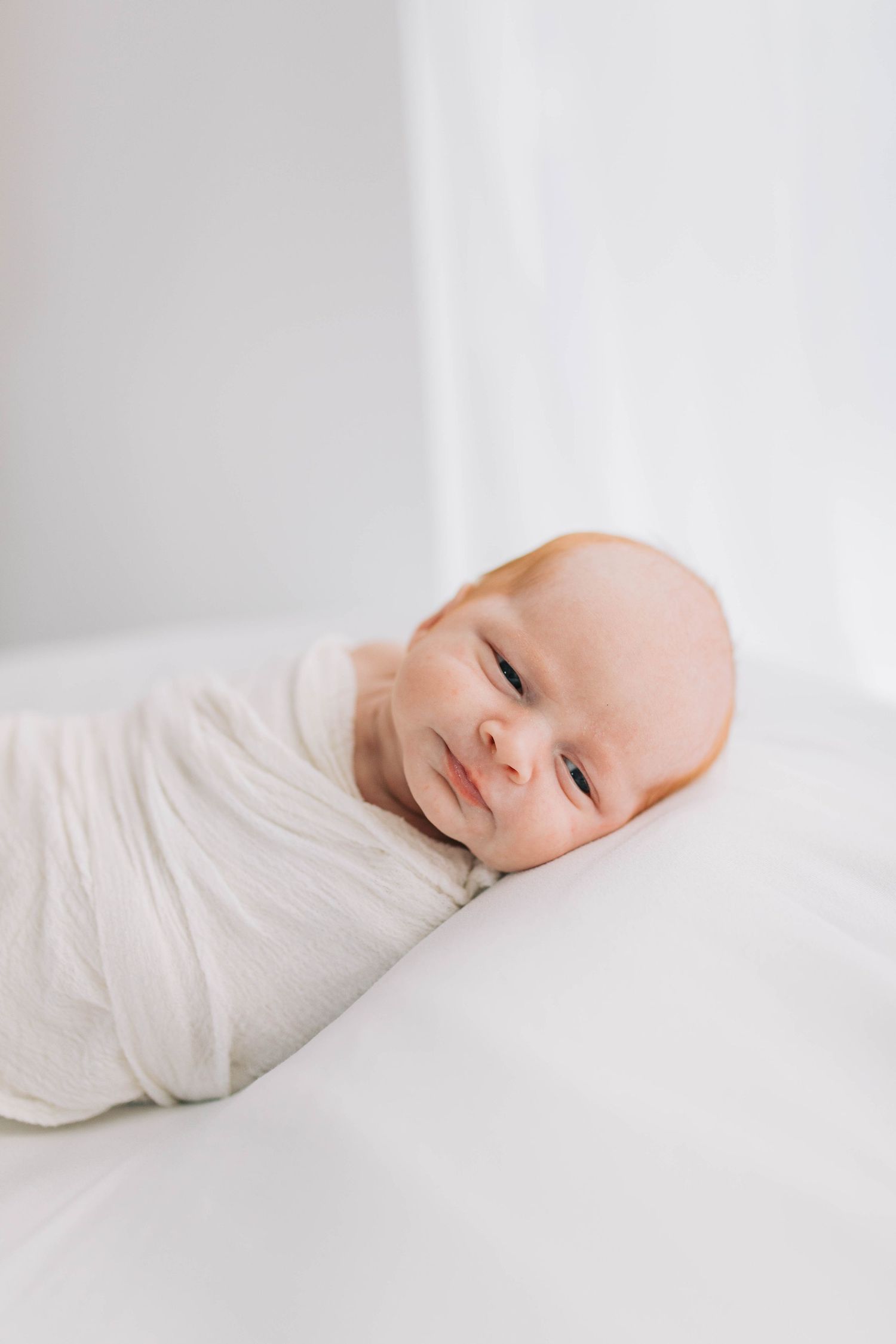 Newborn baby wrapped in white swaddle on white background with gentle smile.