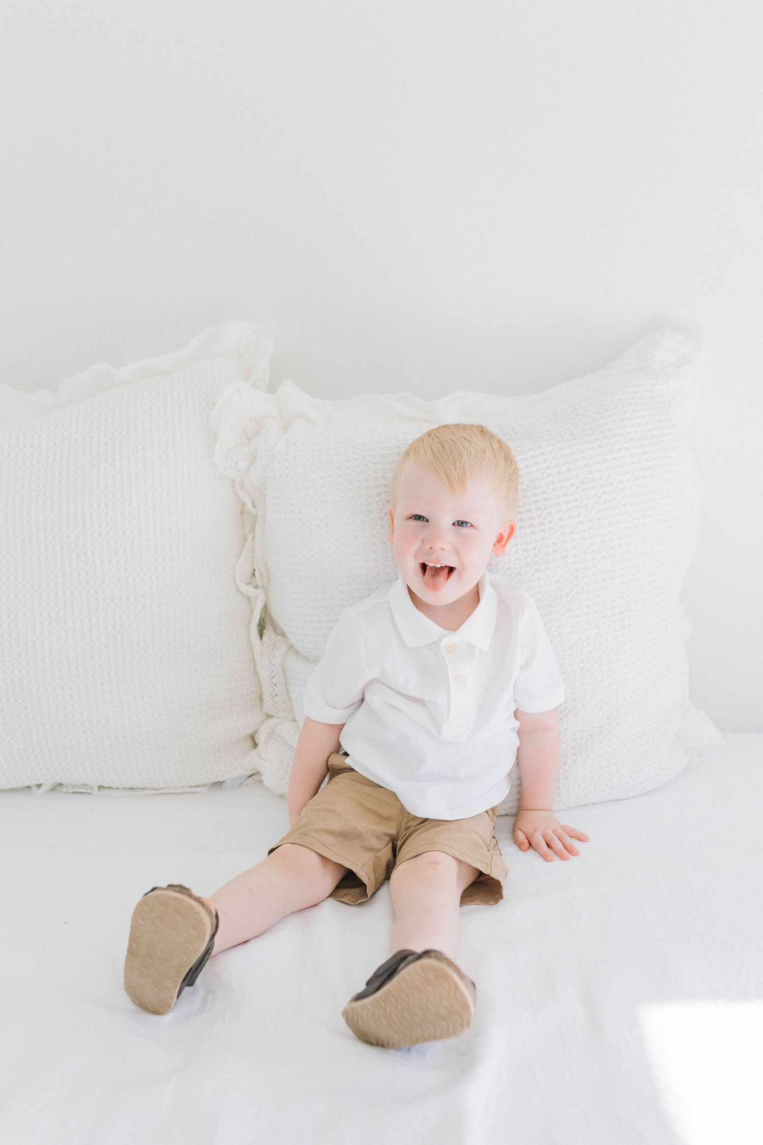 Baby in white shirt and khaki pants smiling while sitting on white bedding.