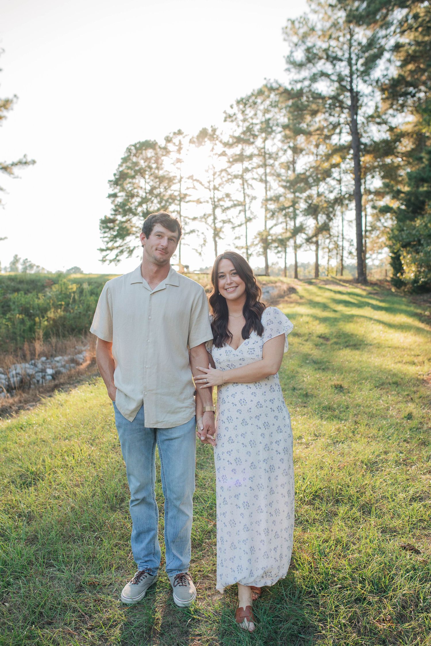 Couple standing together on grassy slope with pine trees during golden hour.
