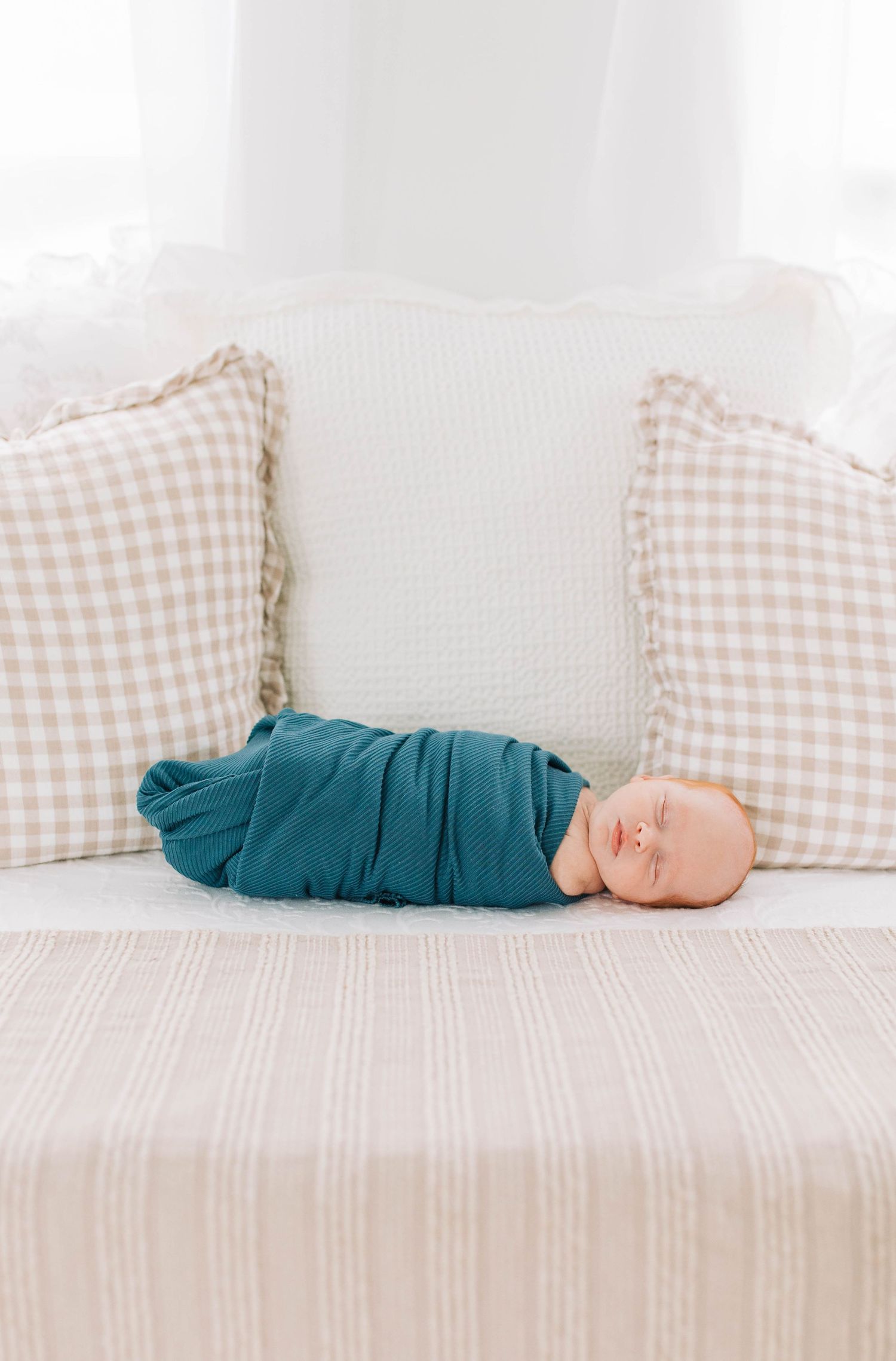 Baby in teal outfit lying on white bed with checkered pillows.