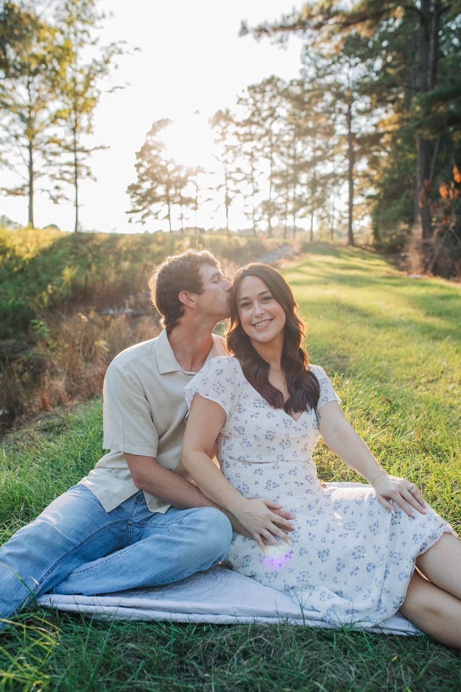 Couple relaxing on blanket in grassy field with pine trees at sunset.