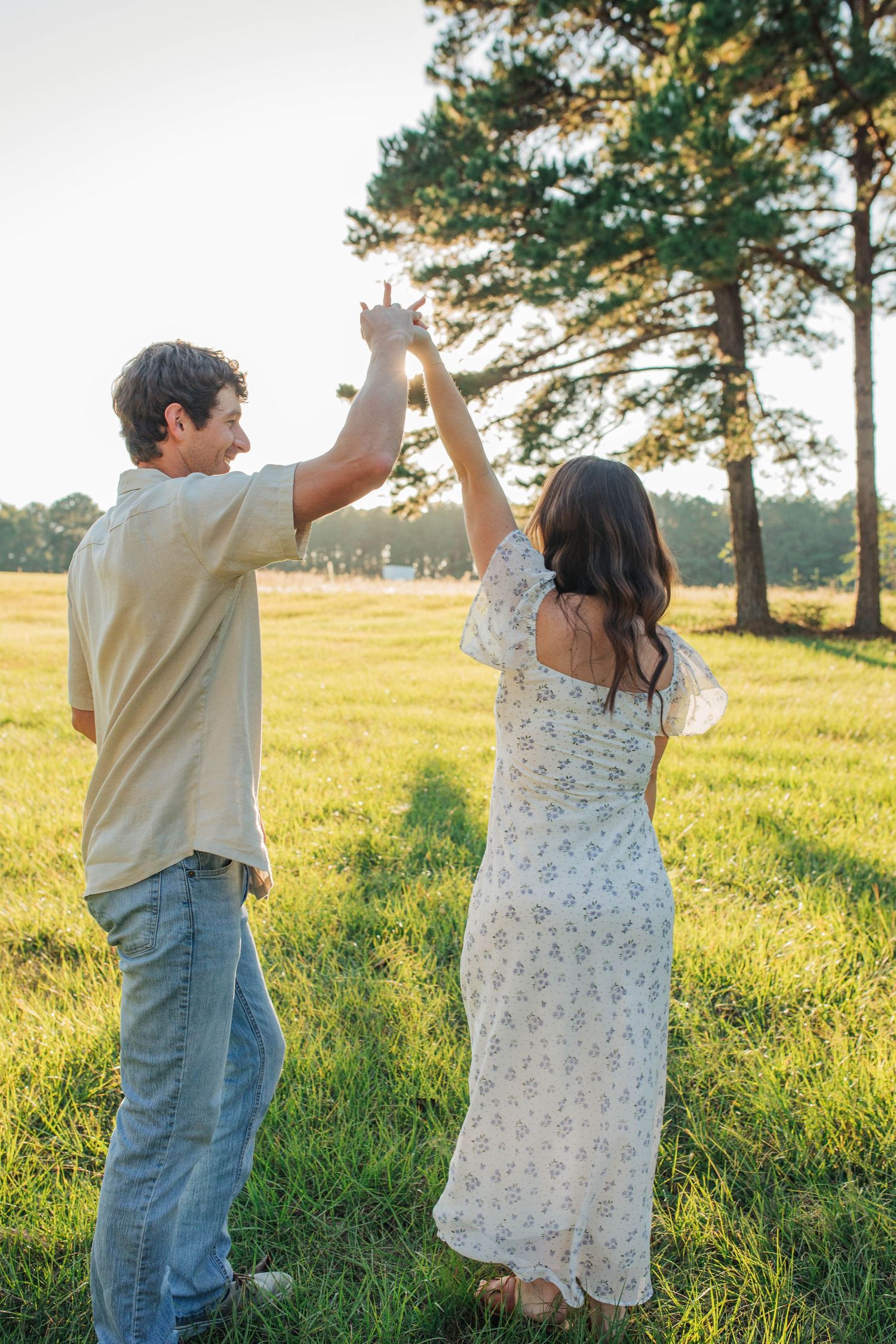Couple dancing together in sunlit field with pine trees.
