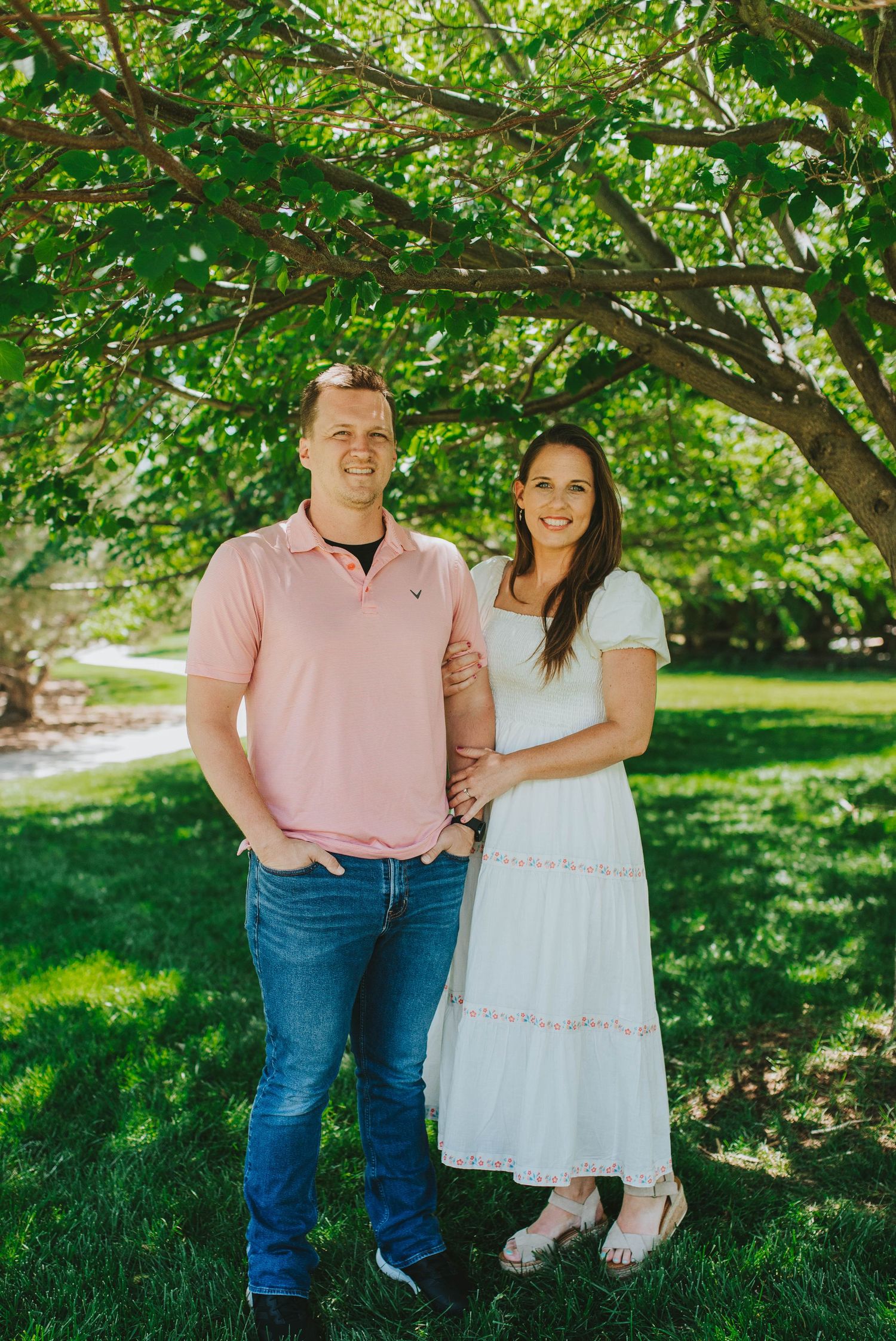 Couple poses together under green trees in a sunny park setting.
