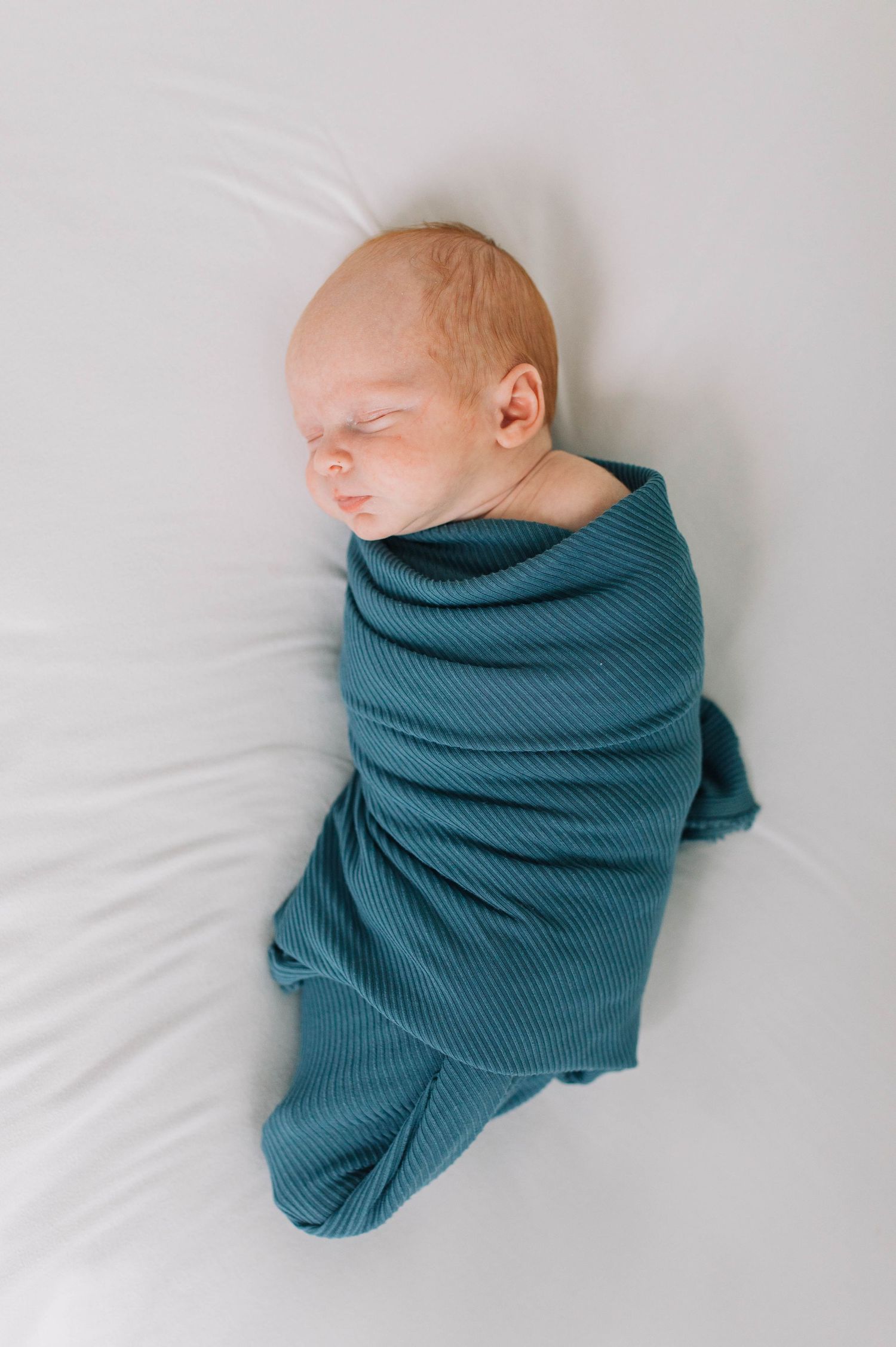 Sleeping infant wrapped in teal ribbed blanket on white bedding.