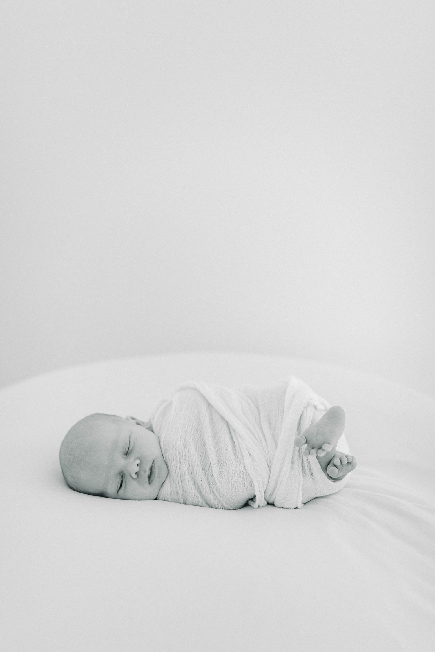 Newborn baby wrapped in white swaddle lying peacefully on white background.