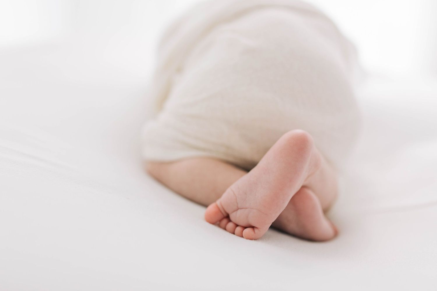 Tiny baby feet peeking out from white swaddle against light background.