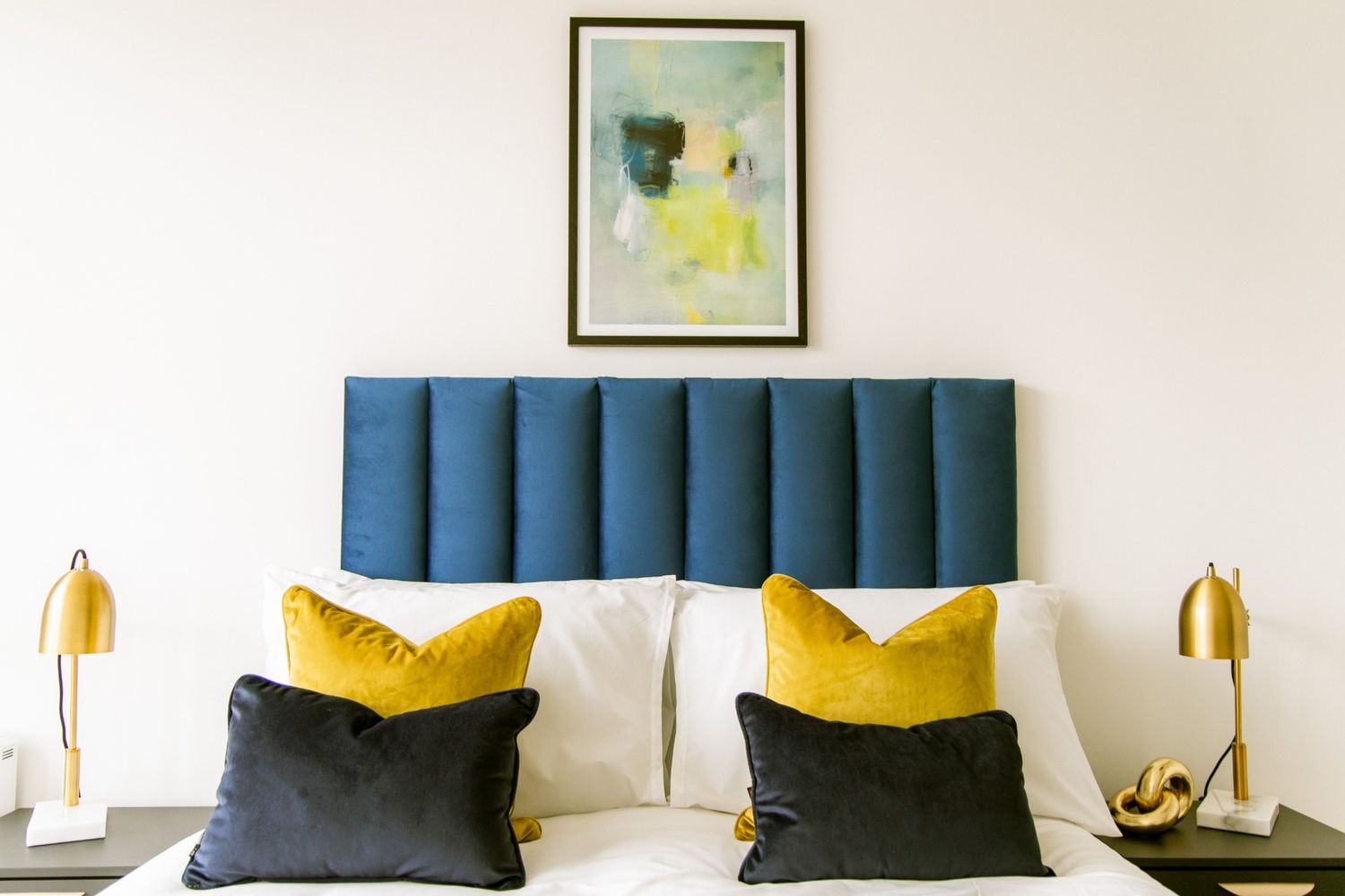 Bold bedroom scene with deep blue headboard, mustard yellow and navy pillows, and golden bedside lamps — styled for modern living.