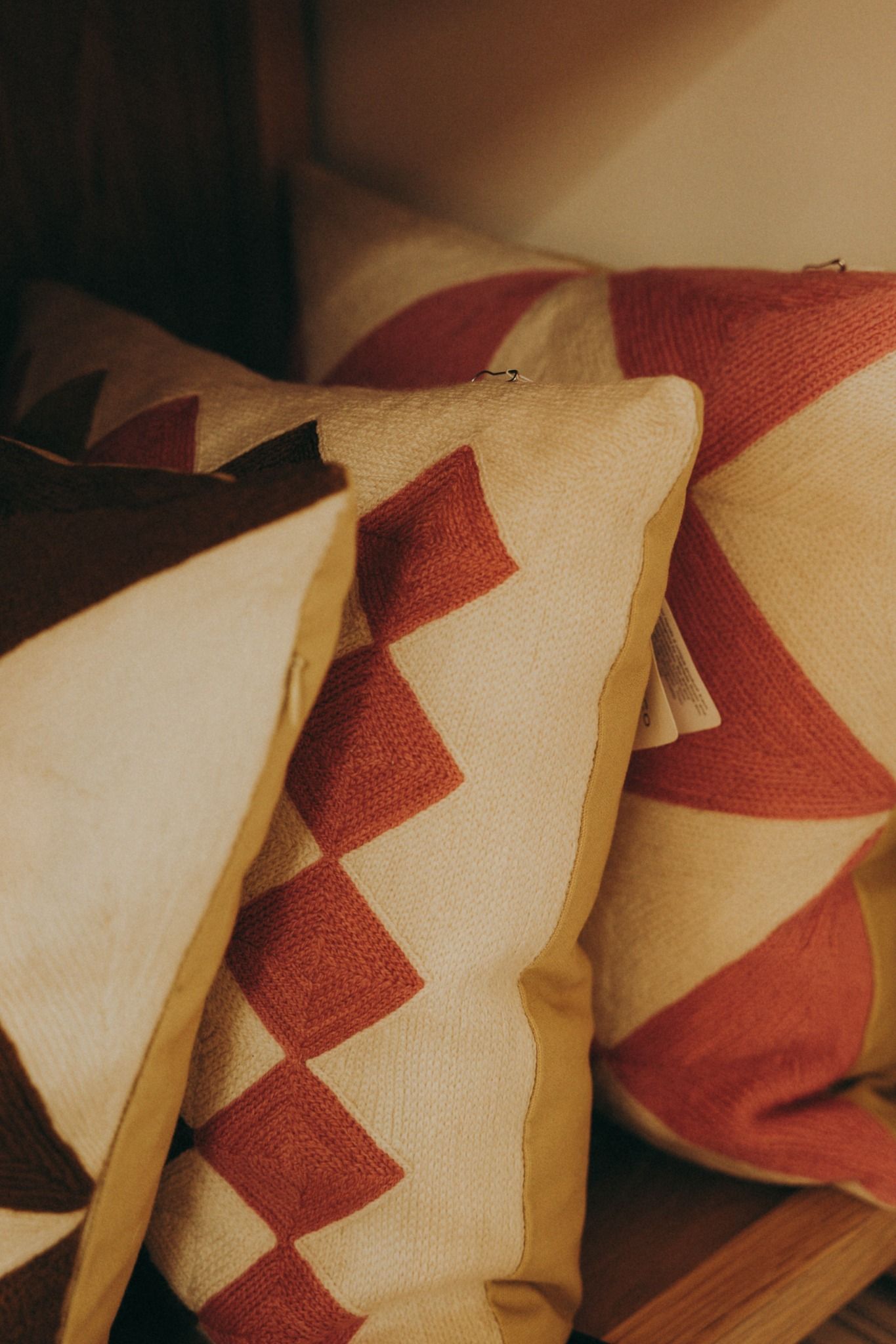 Close-up of bold geometric patterned cushions in red and brown tones, nestled on wooden shelves — styled with a warm aesthetic by Sigi Photo