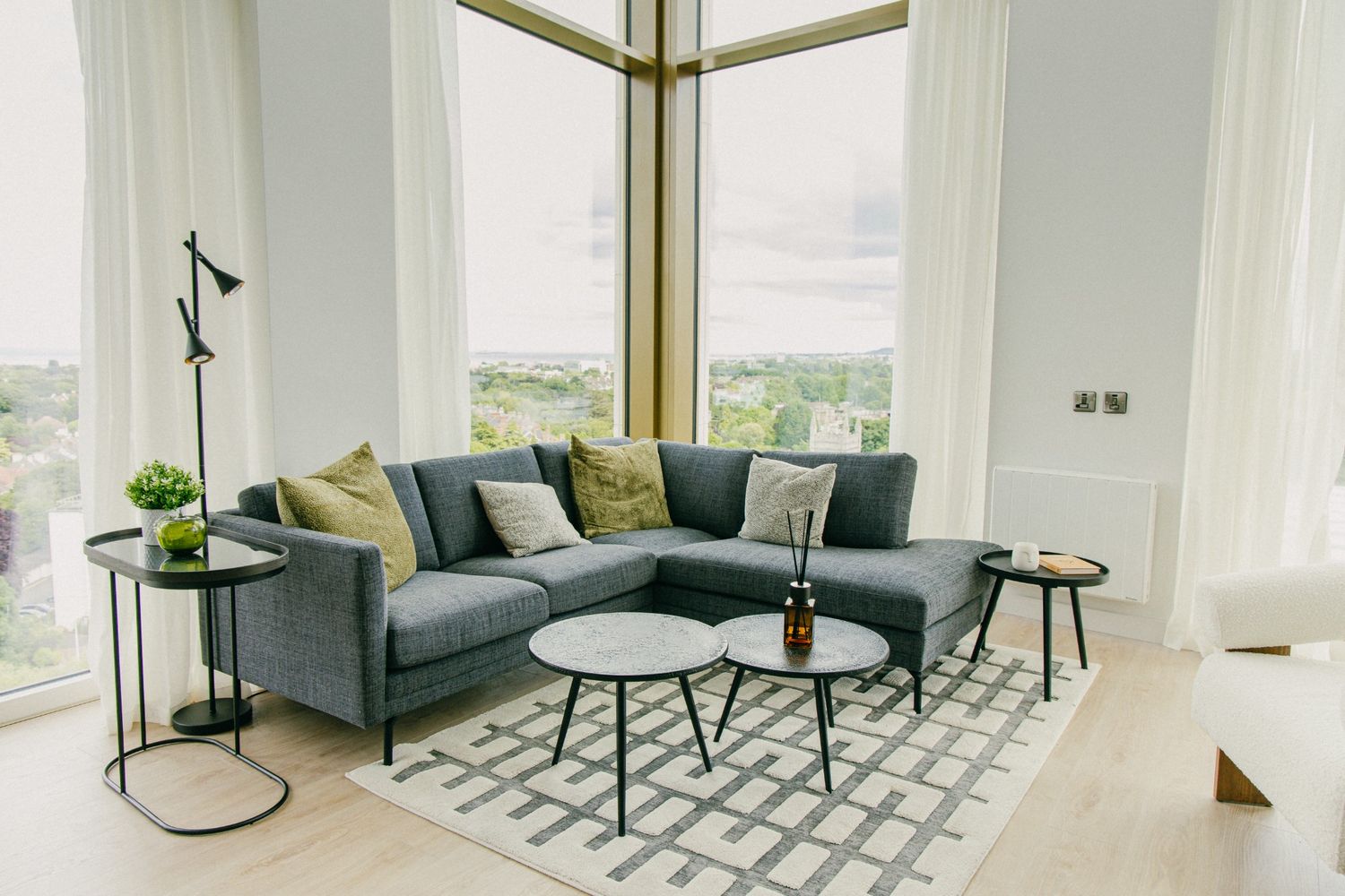 Spacious modern living room with grey sectional sofa, textured rug, and panoramic city views through tall windows.