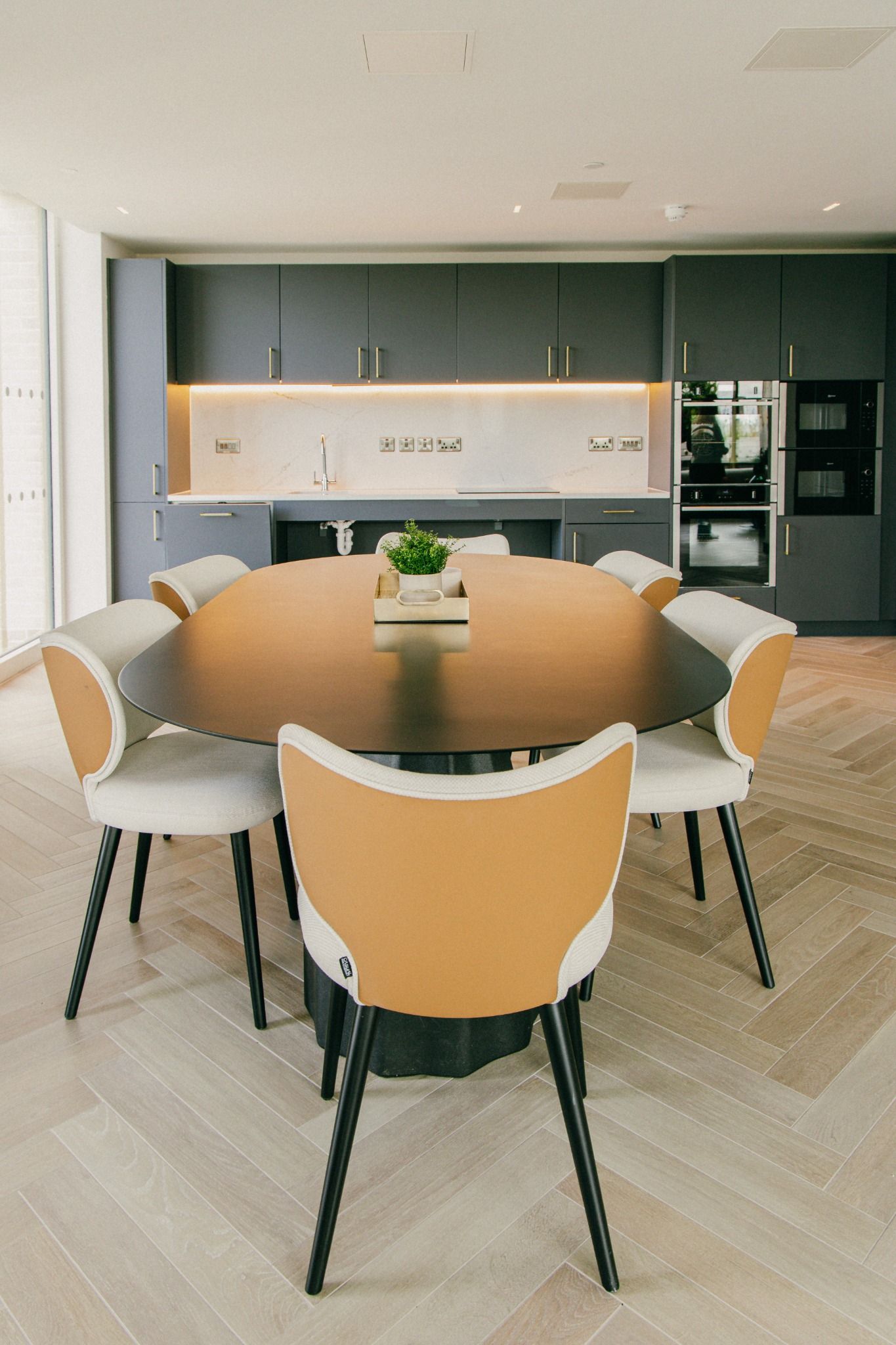Modern kitchen with dark cabinetry and oval dining table surrounded by stylish camel and cream chairs, set on a herringbone floor in Dublin