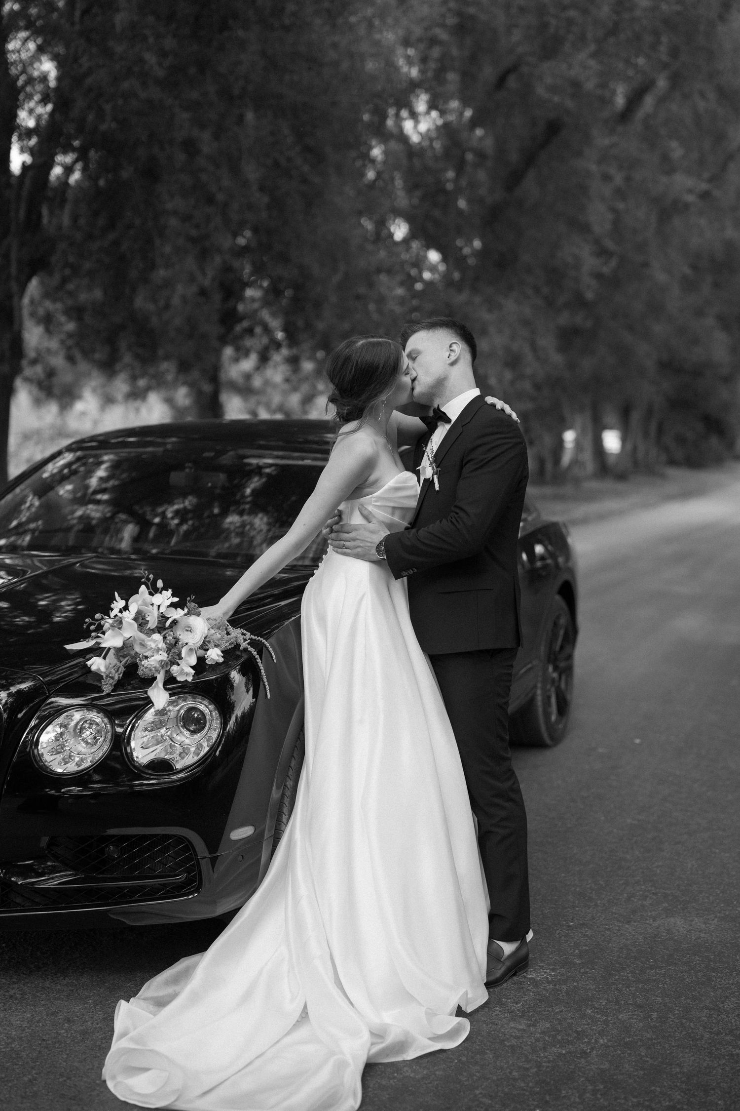 Romantic black and white wedding photo of couple by luxury car with bride's dress trailing.