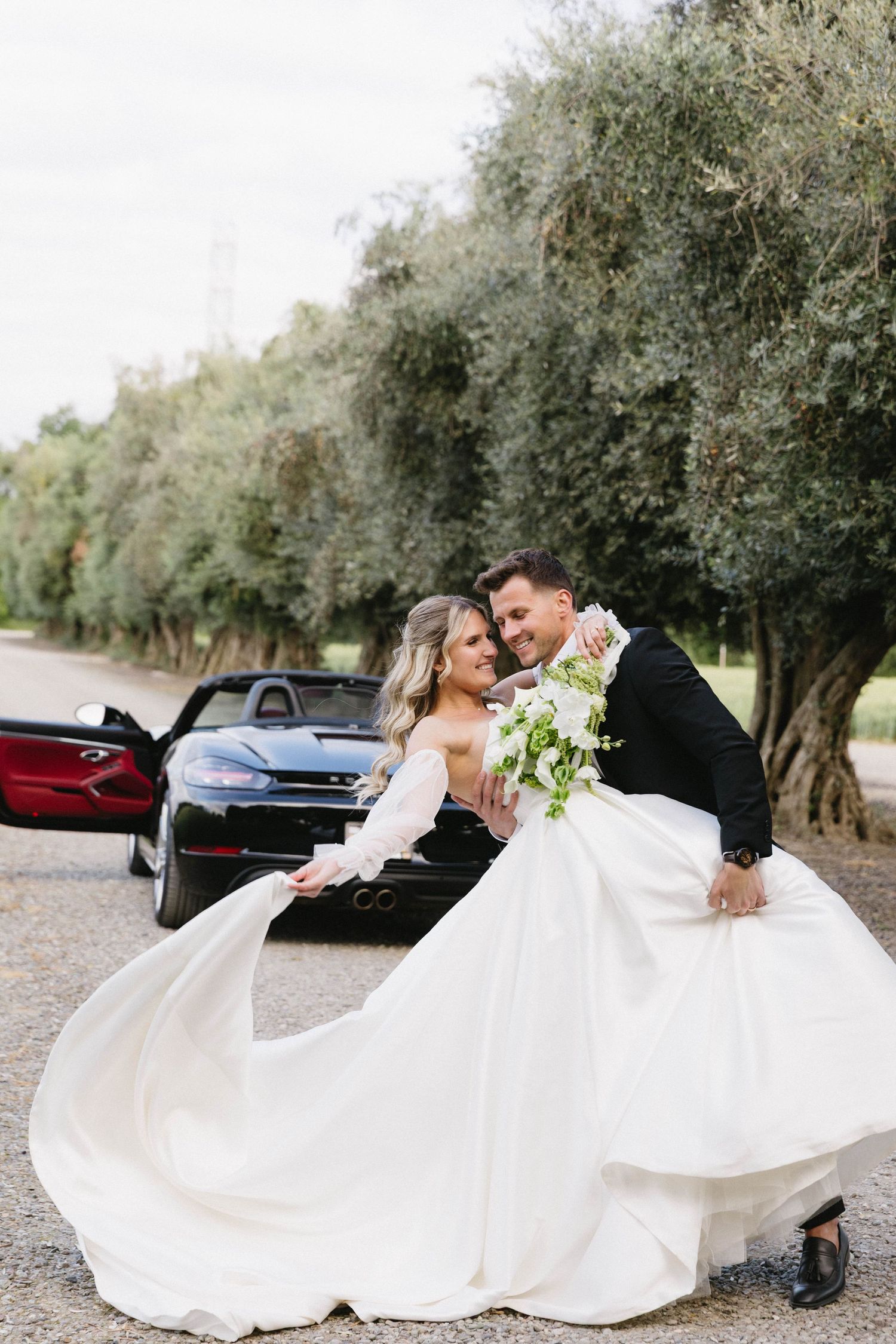 Elegant wedding portraits with flowing white dress and black sports car among olive trees.