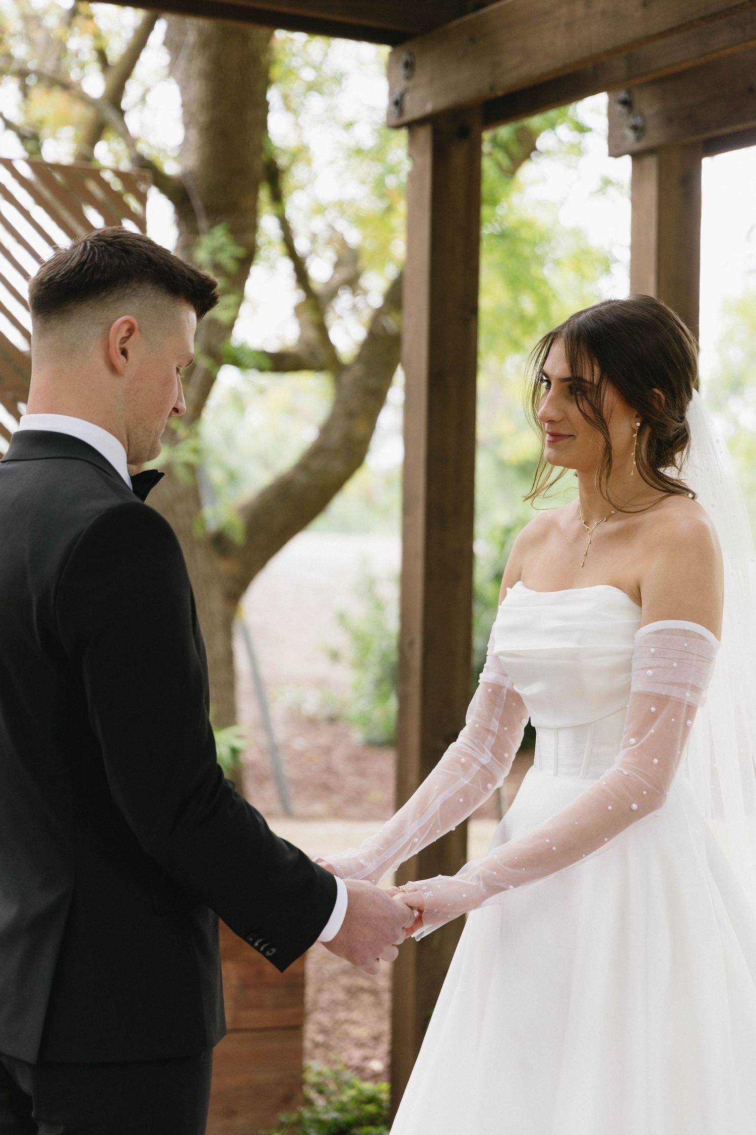 Couple holding hands during intimate wedding ceremony under wooden pergola.