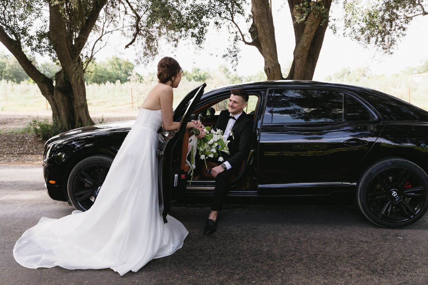 Wedding couple posing with a sleek black luxury car under a canopy of trees.