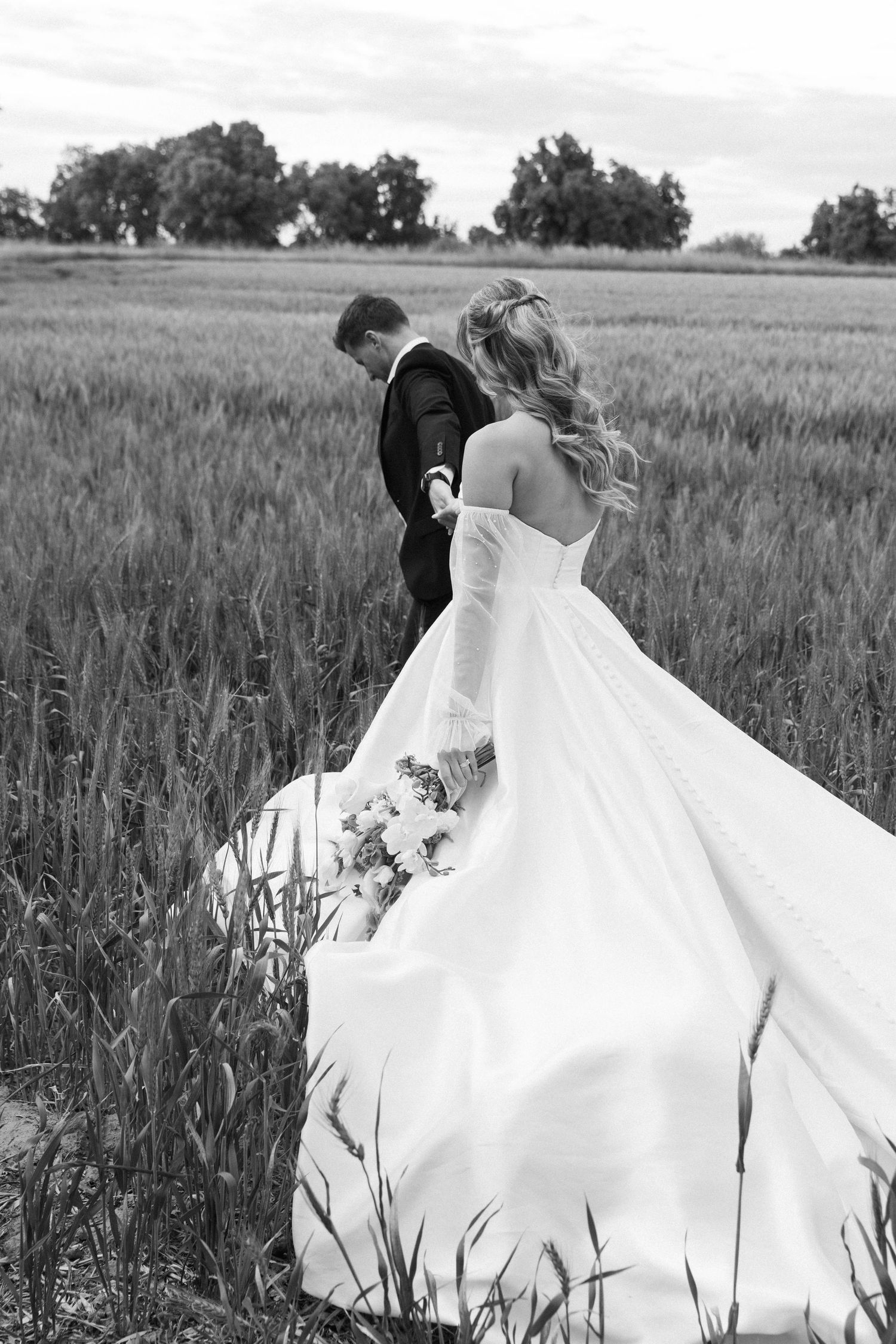 Black and white photo of couple walking through wheat field with flowing wedding dress.