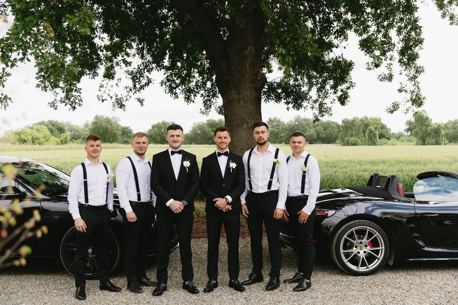 Wedding party members in black and white attire gathered around luxury vehicles for a group photo.