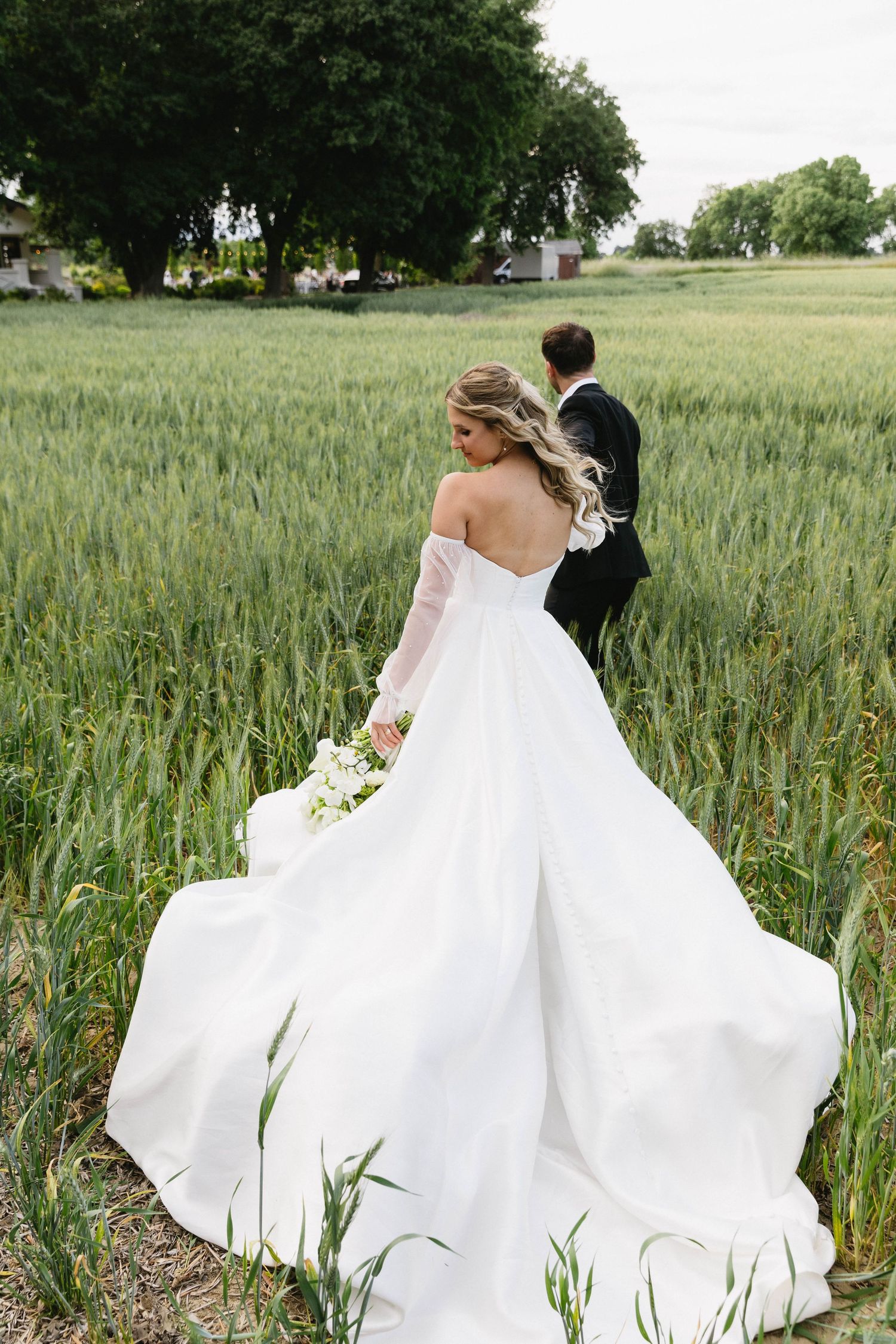 Wedding couple walking through green wheat field with flowing white dress train.
