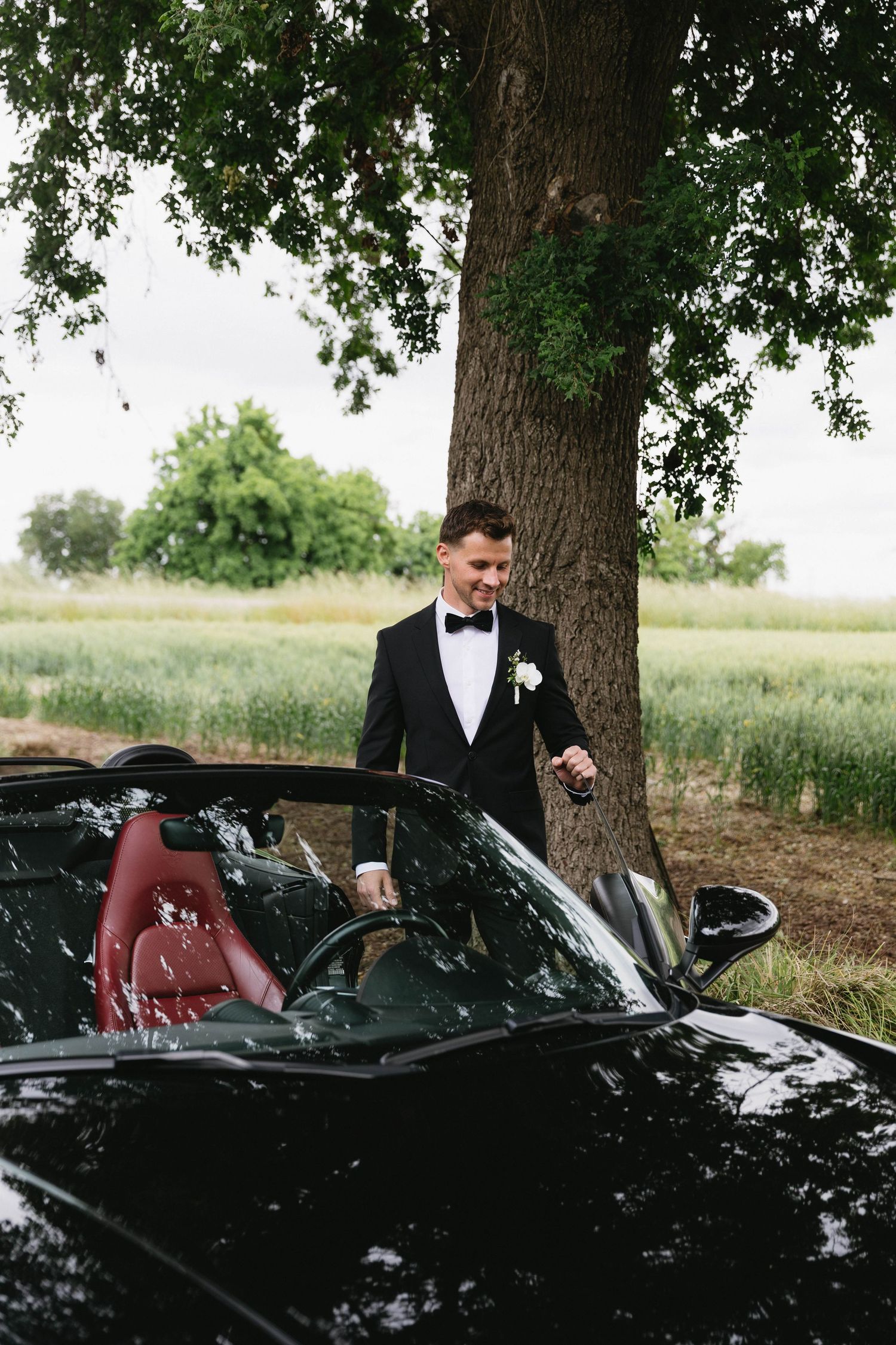 A sleek black convertible car parked under trees with someone in a black tuxedo sitting in the driver's seat.