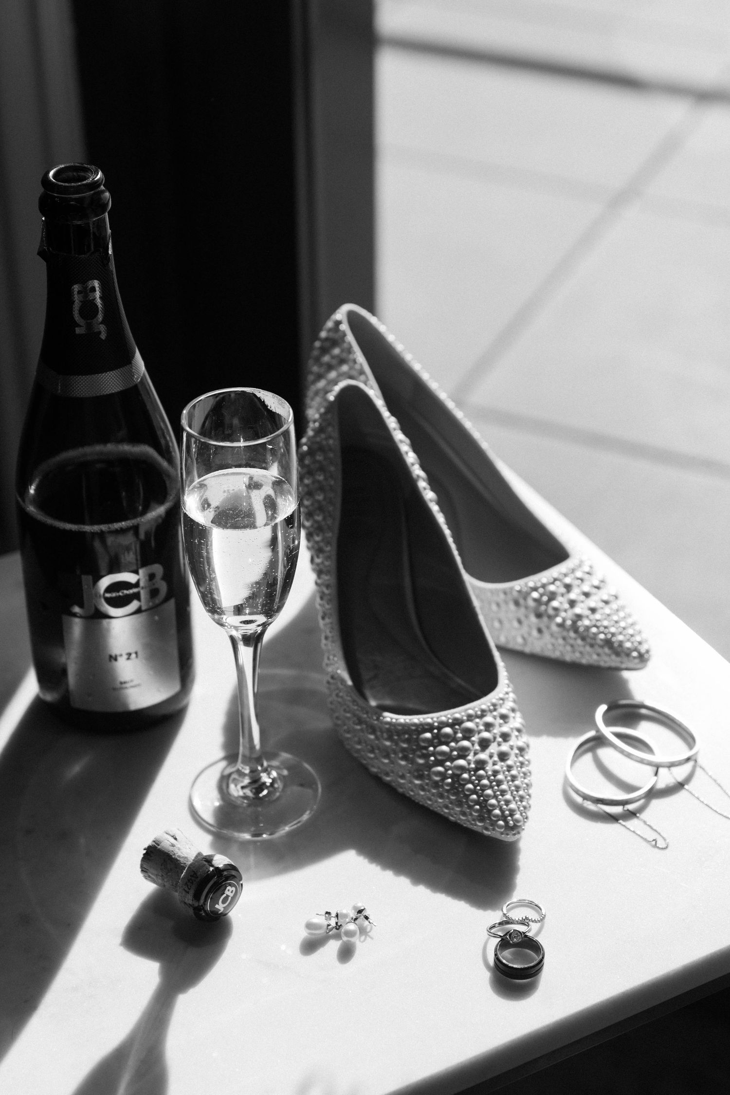 Black and white photo of wedding accessories including champagne, studded bridal shoes, rings and jewelry on a table.