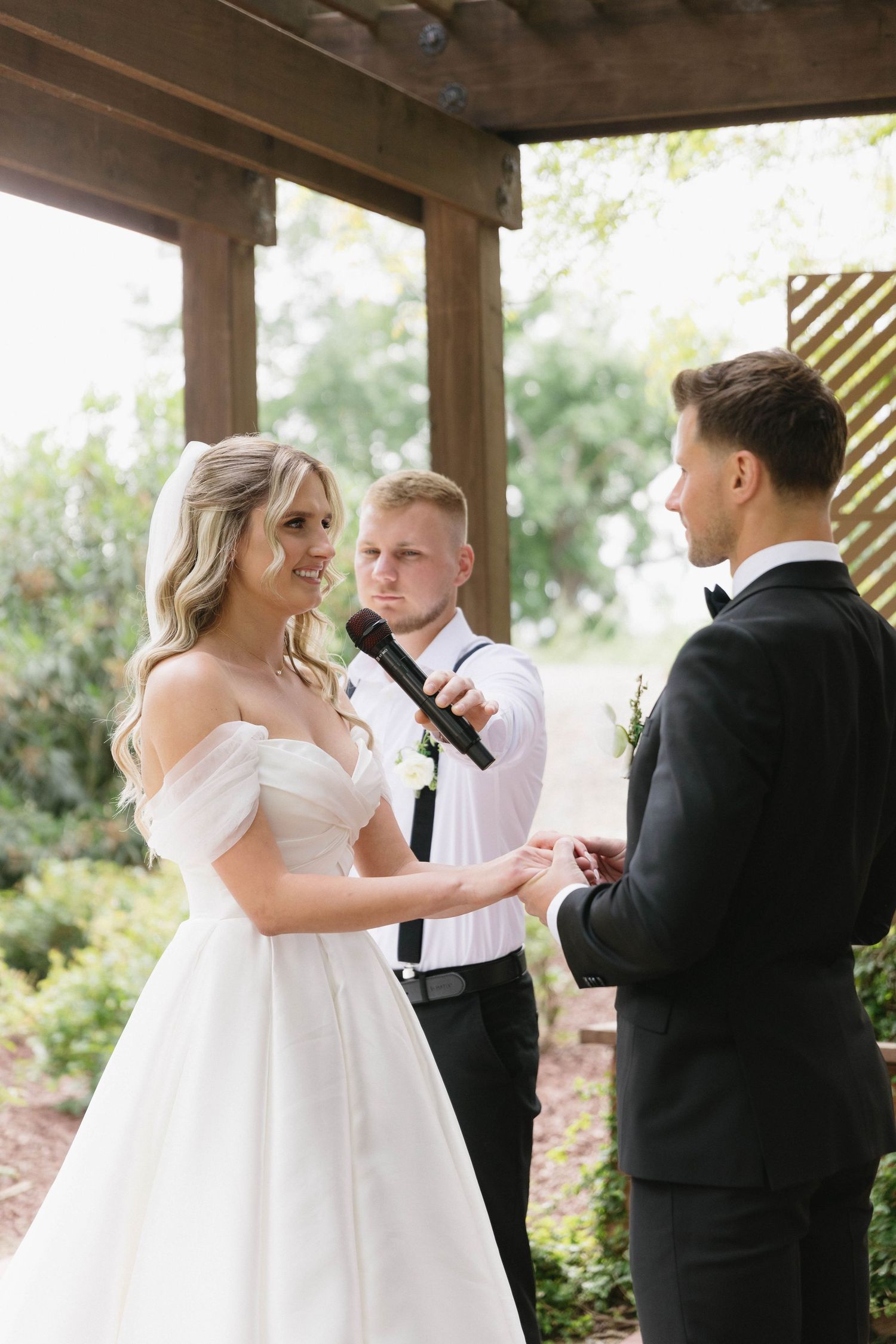 Wedding ceremony under wooden pergola with couple exchanging vows and officiant.