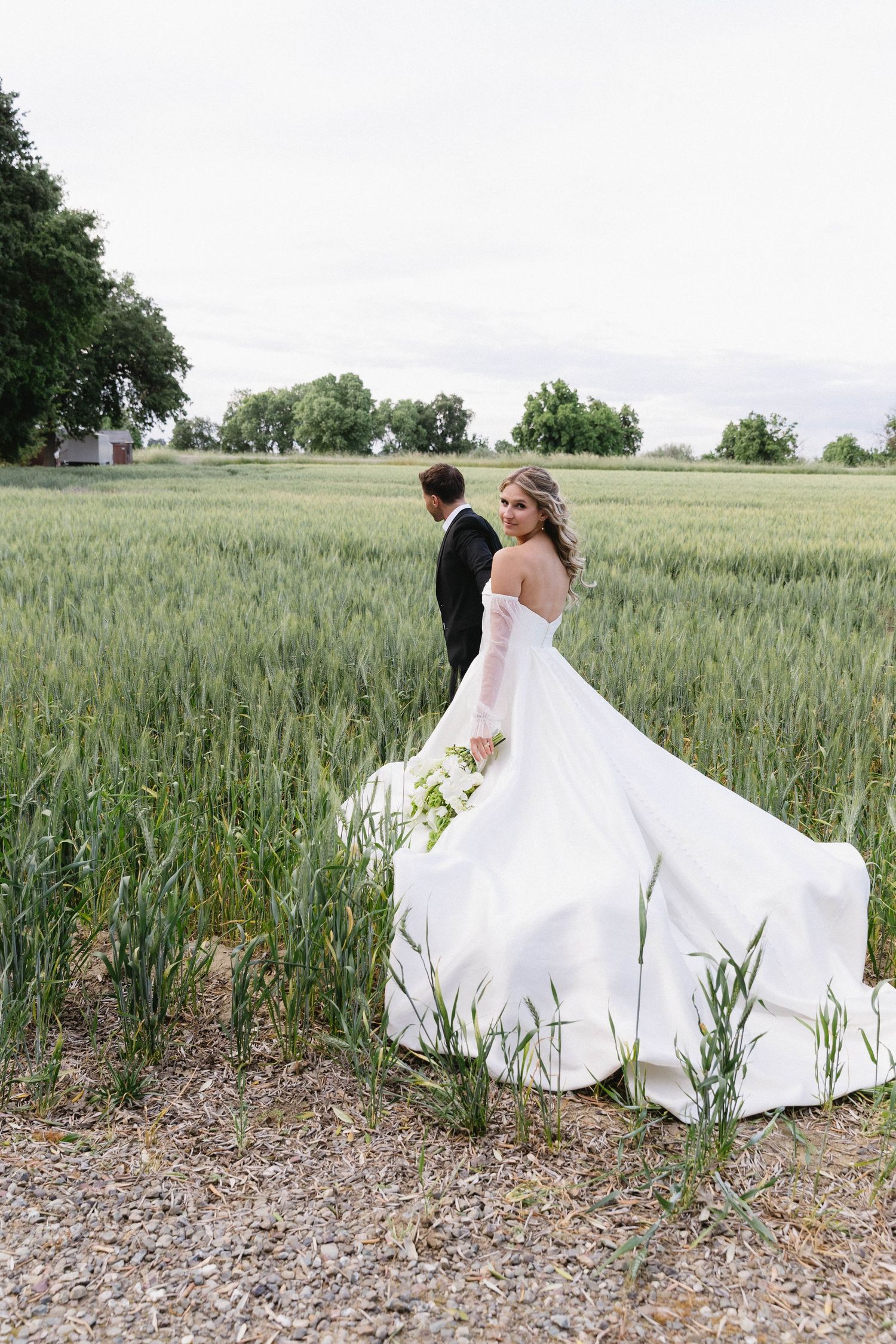 A bride's flowing white dress trails behind her in a scenic wheat field.