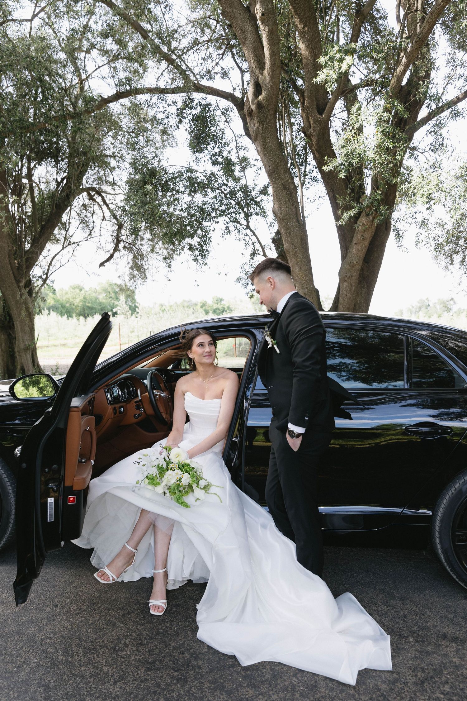 Bride and groom pose playfully by black luxury car under trees.