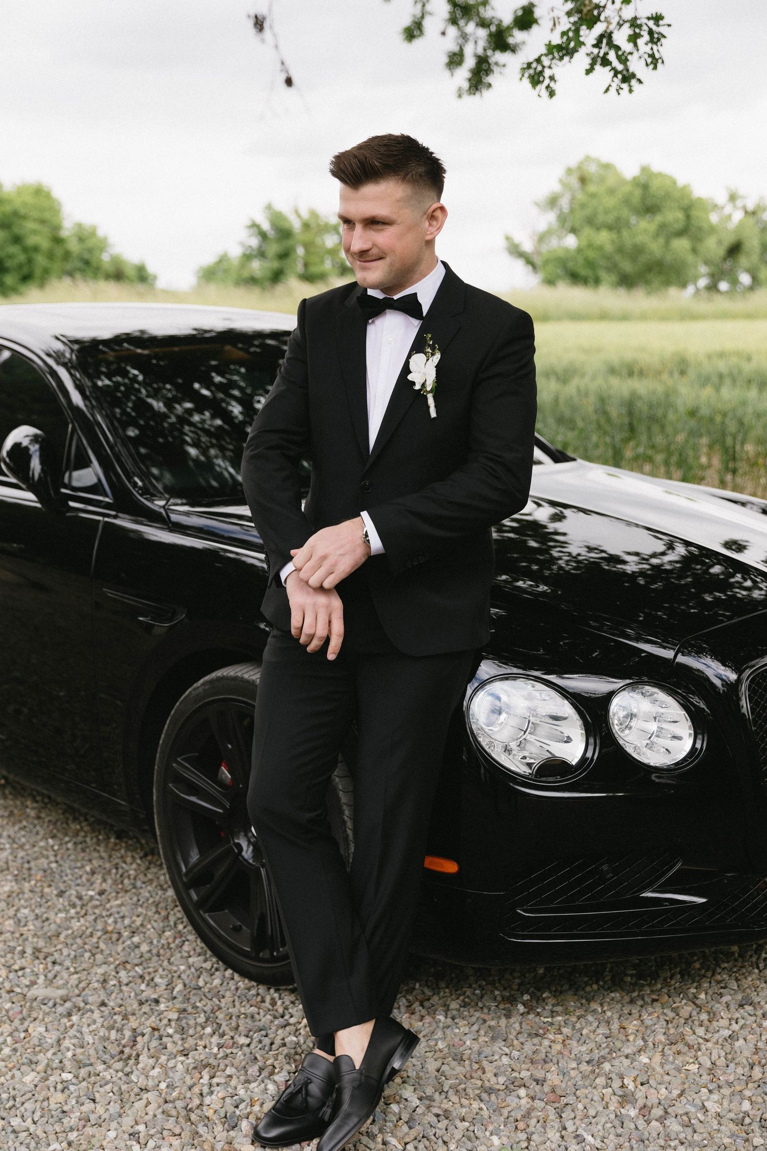 Groom in formal attire leaning against black luxury car during wedding photoshoot.