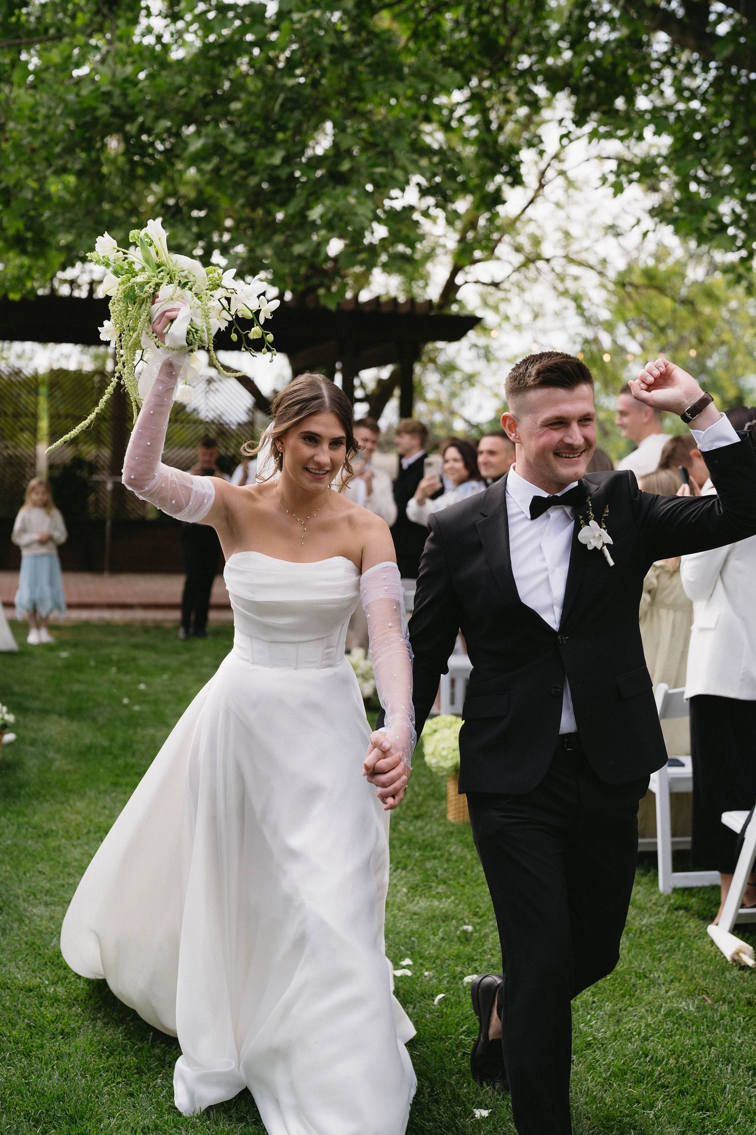 Joyful wedding celebration with dancing and raised arms on a sunny lawn.