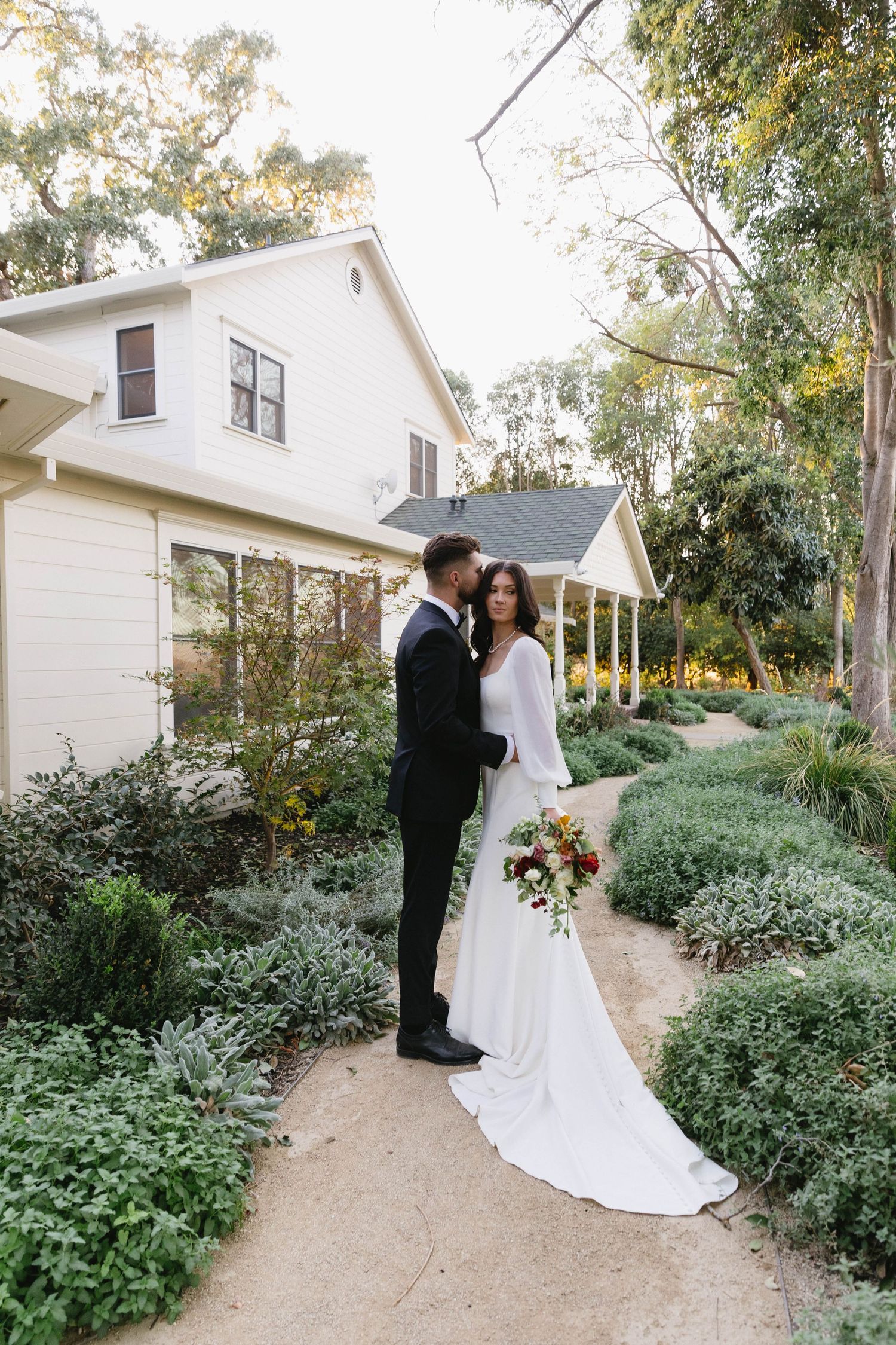 A bride and groom share an intimate moment on a garden path surrounded by lush greenery and white cottages.