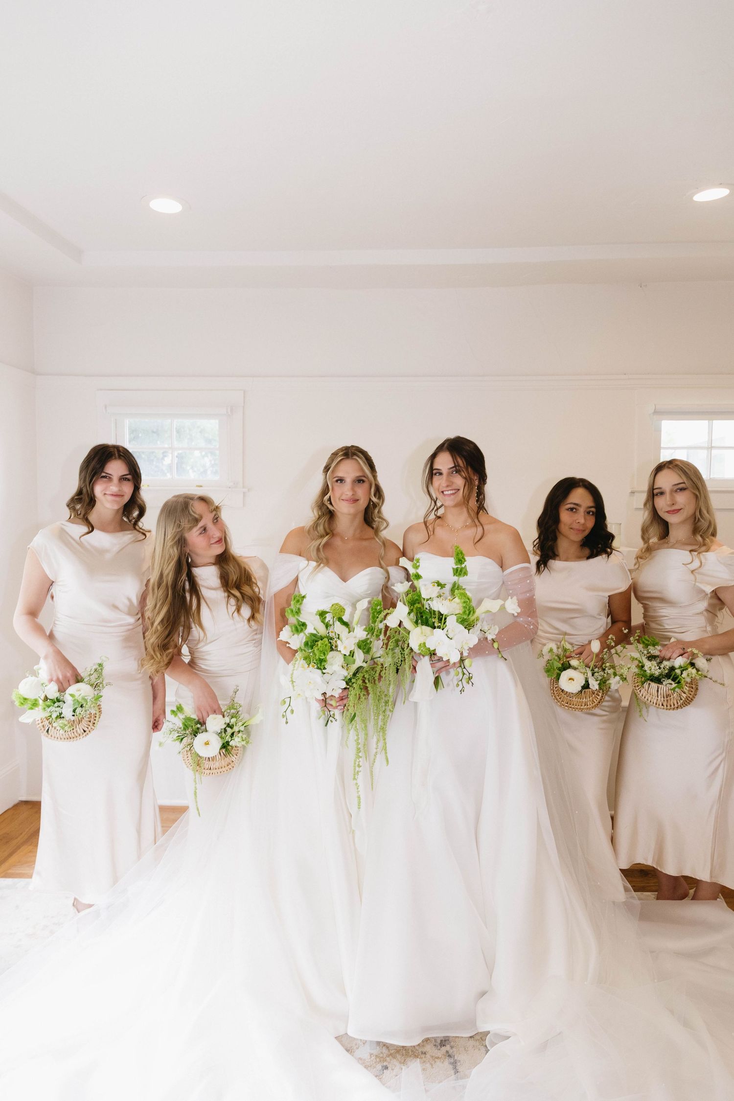 Bridesmaids in matching white gowns pose together holding cascading green and white bouquets.