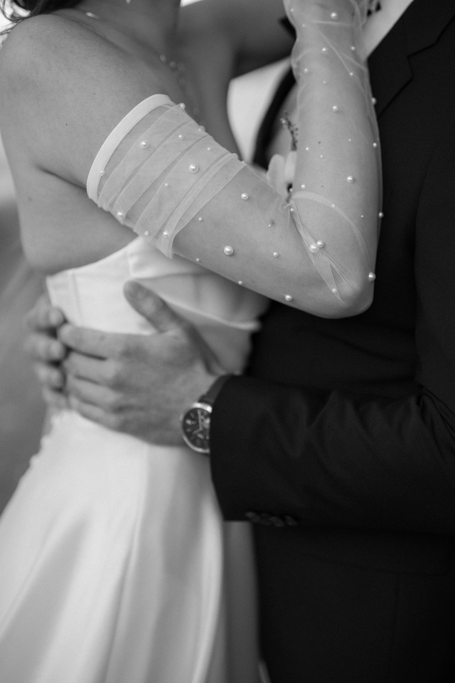 Close-up detail of bride's pearl-dotted sheer sleeve against groom's black tuxedo jacket.