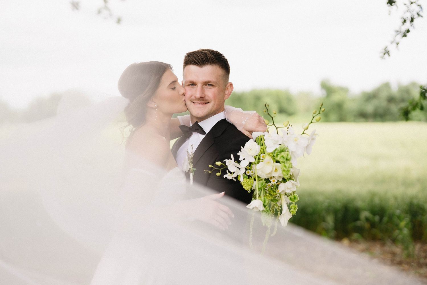 A romantic wedding couple embraces in a sunlit meadow while holding a bouquet of white and green flowers.