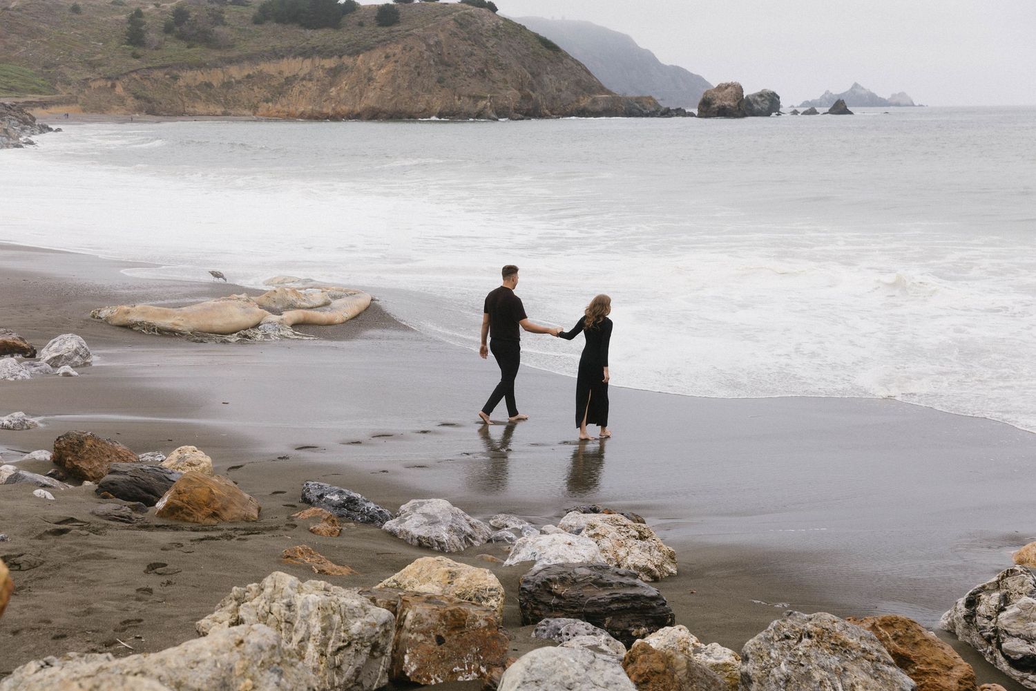 Two silhouettes stand together on rocky beach with misty coastal cliffs and crashing waves in the background.