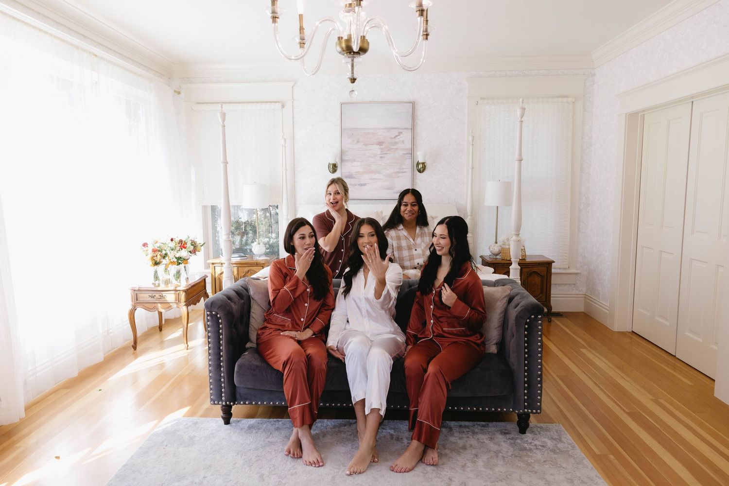 Wedding party in rust-colored dresses relaxing in an elegant white room with chandelier.