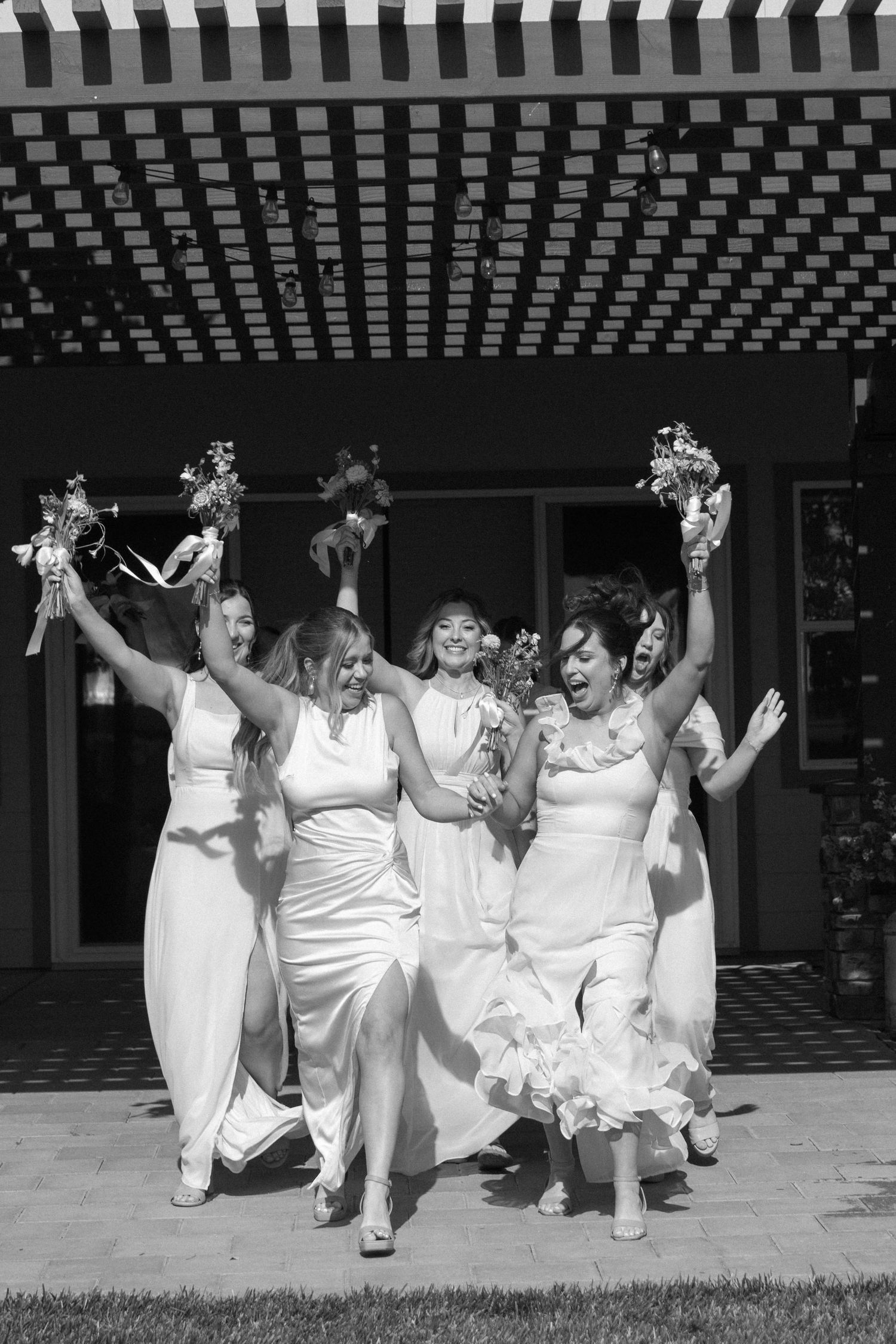 Bridesmaids in white dresses celebrating joyfully under a striped pergola.