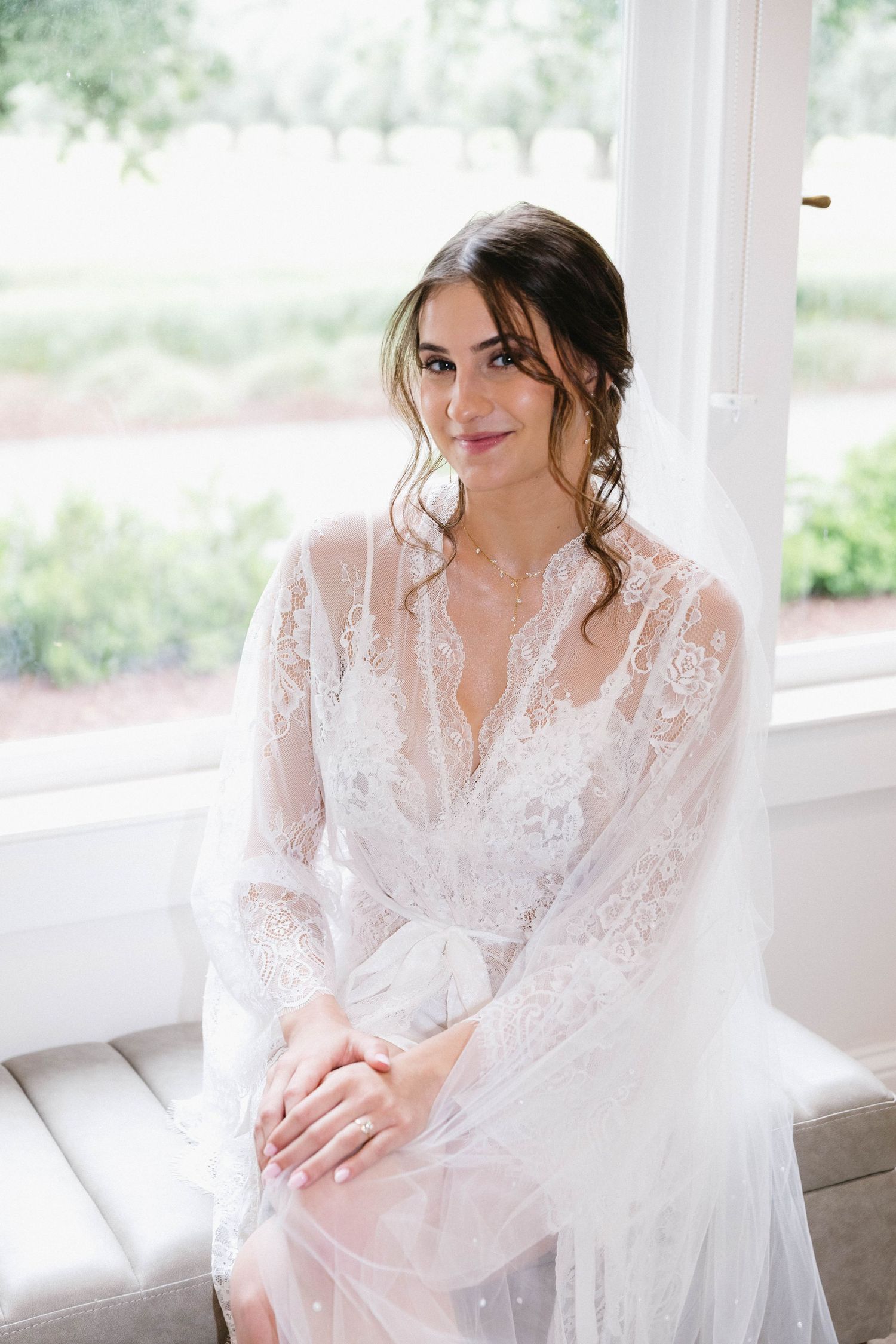 A bride in a delicate lace robe poses by window light during wedding preparations.