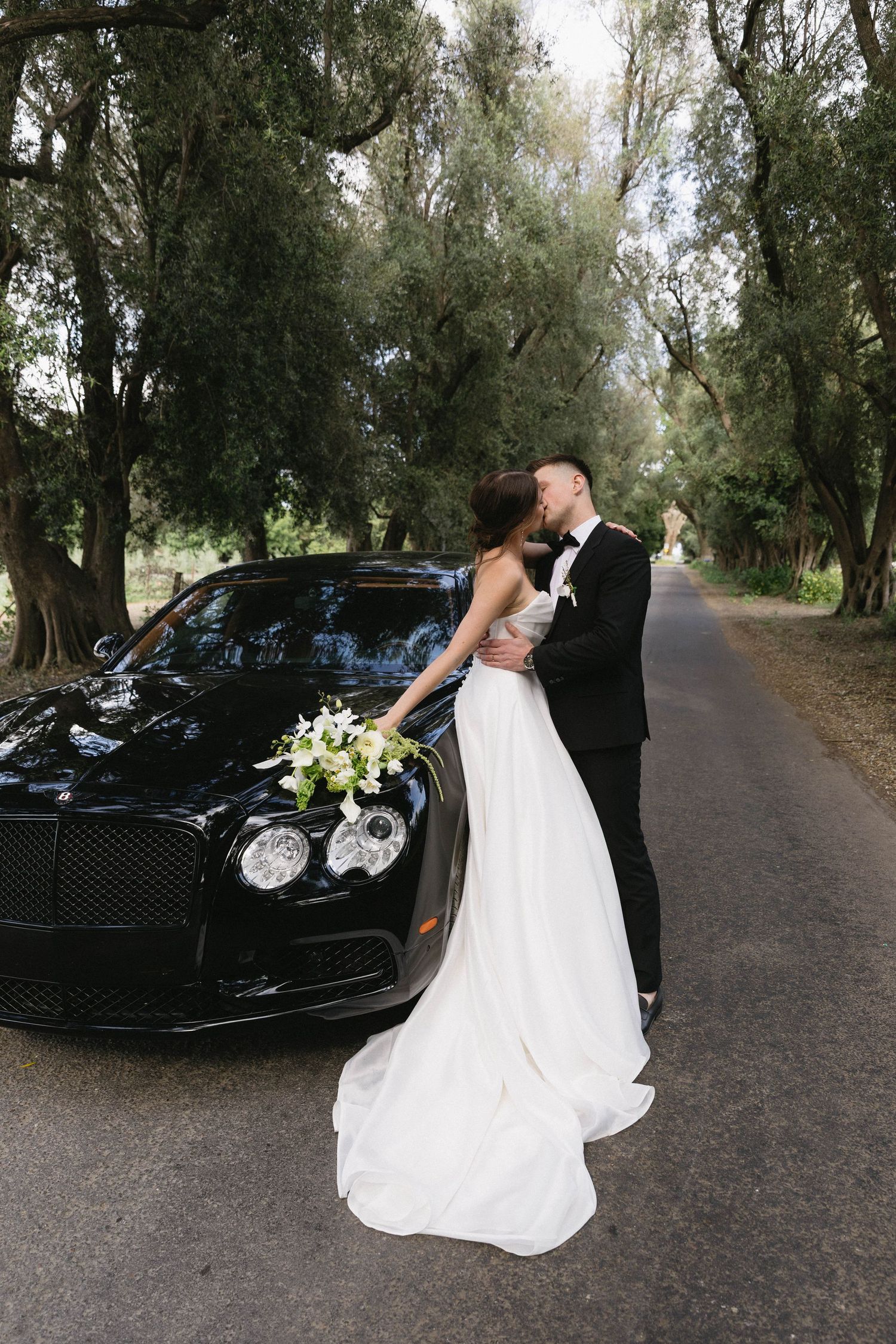 A romantic wedding couple poses with their black luxury car on a tree-lined path during an outdoor photo session.