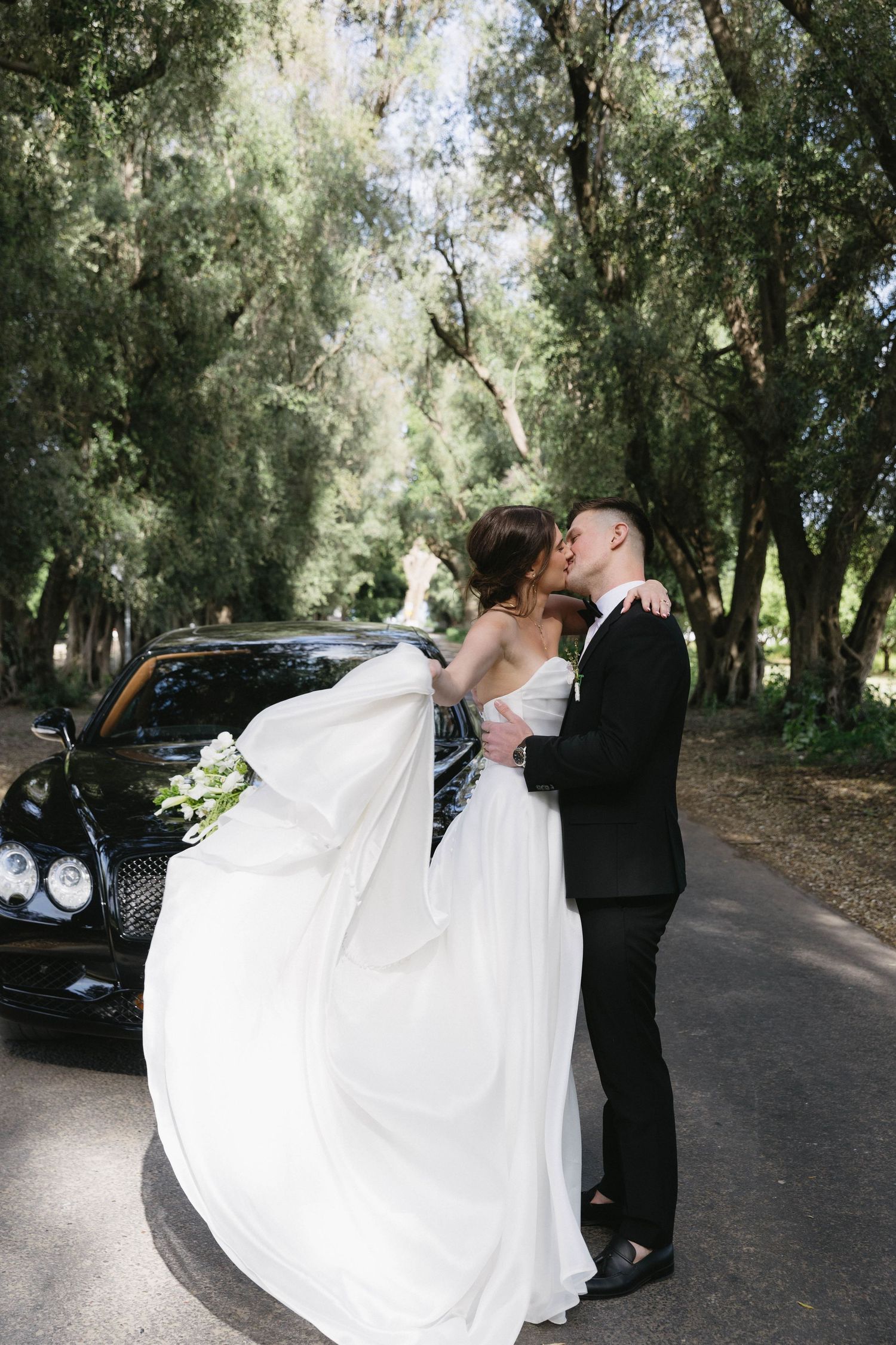Newlyweds share romantic moments on a tree-lined path next to a luxury car.