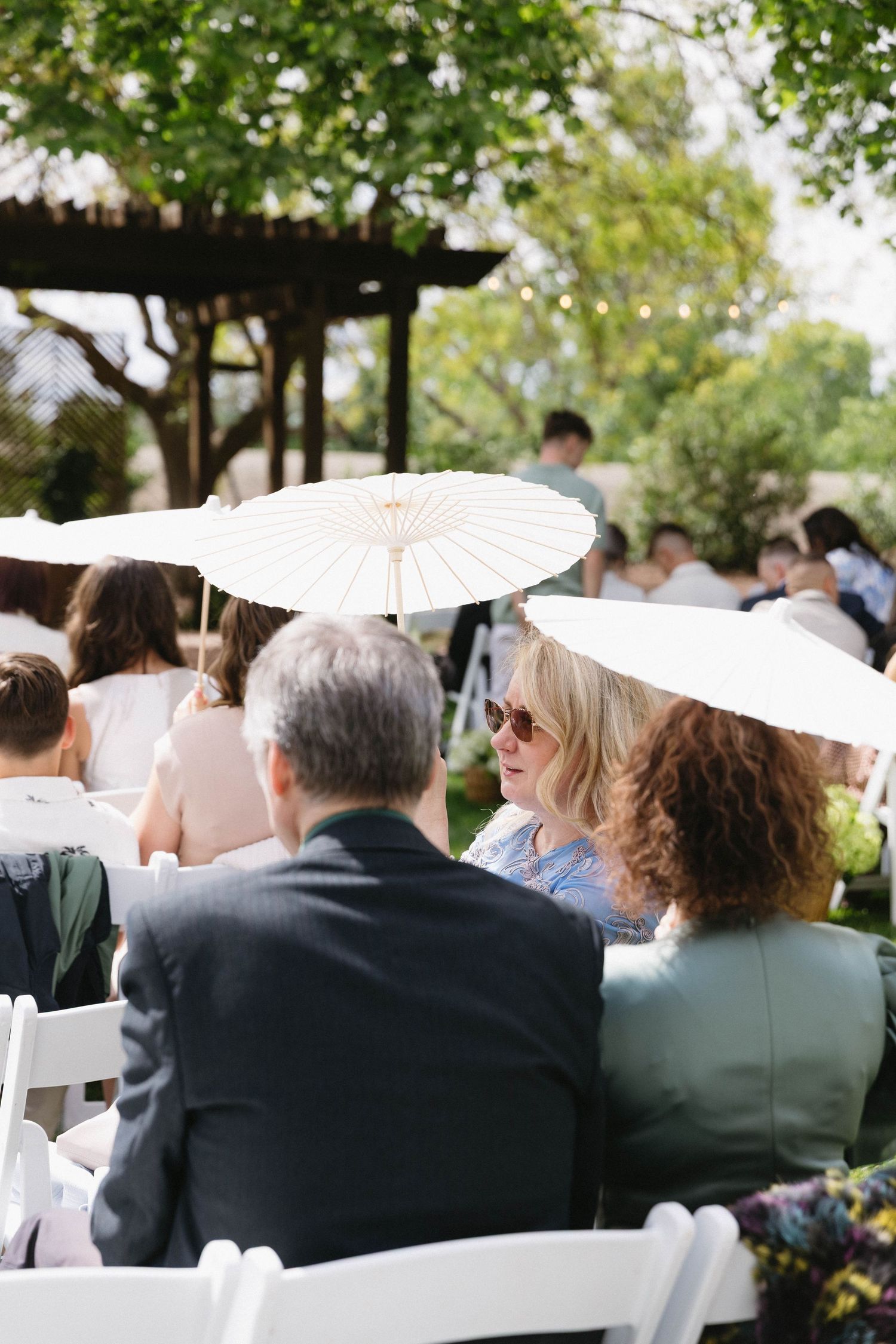 Wedding guests seated in white chairs with parasols during outdoor garden ceremony.