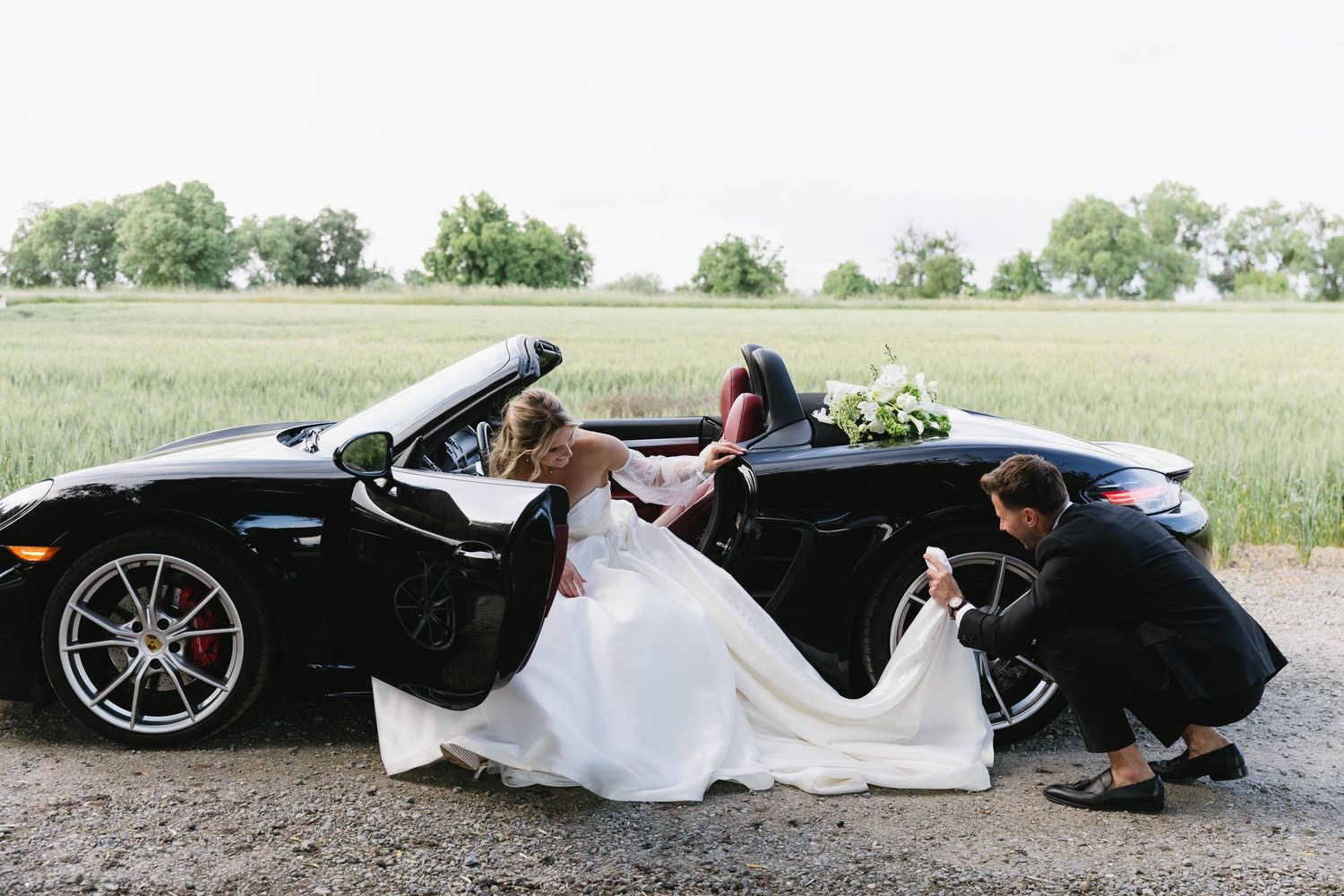 A bride's white wedding dress train flows elegantly beside a sleek black sports car on a country road.