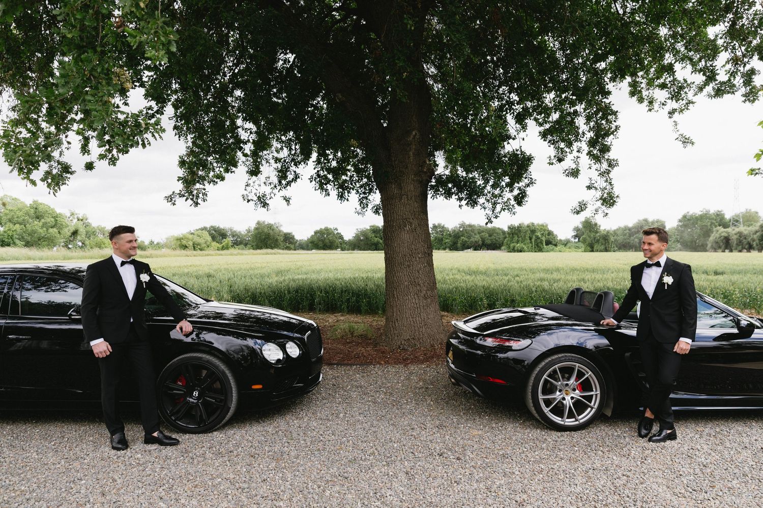 A group of formally dressed people posing with luxury vehicles under trees in an outdoor setting.