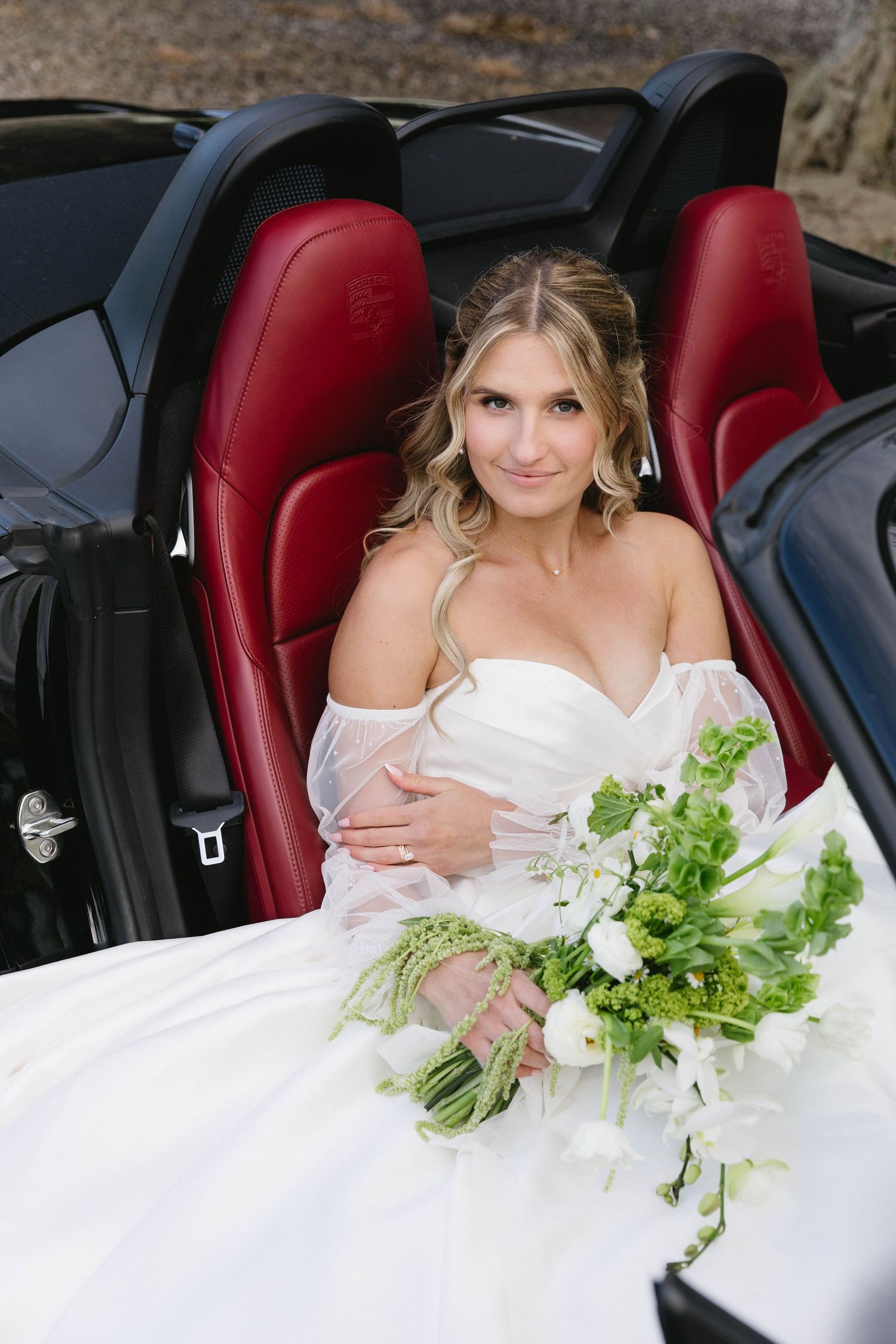 Bride sitting in luxury convertible with red leather seats holding white floral bouquet.