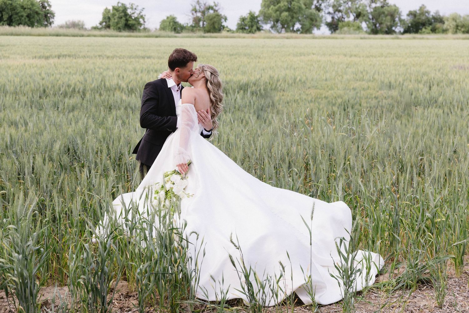 Wedding couple embracing in field with luxury sports car and trees in background.