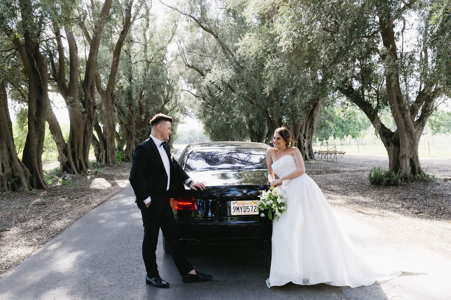 Wedding couple poses on tree-lined path with luxury vehicle.