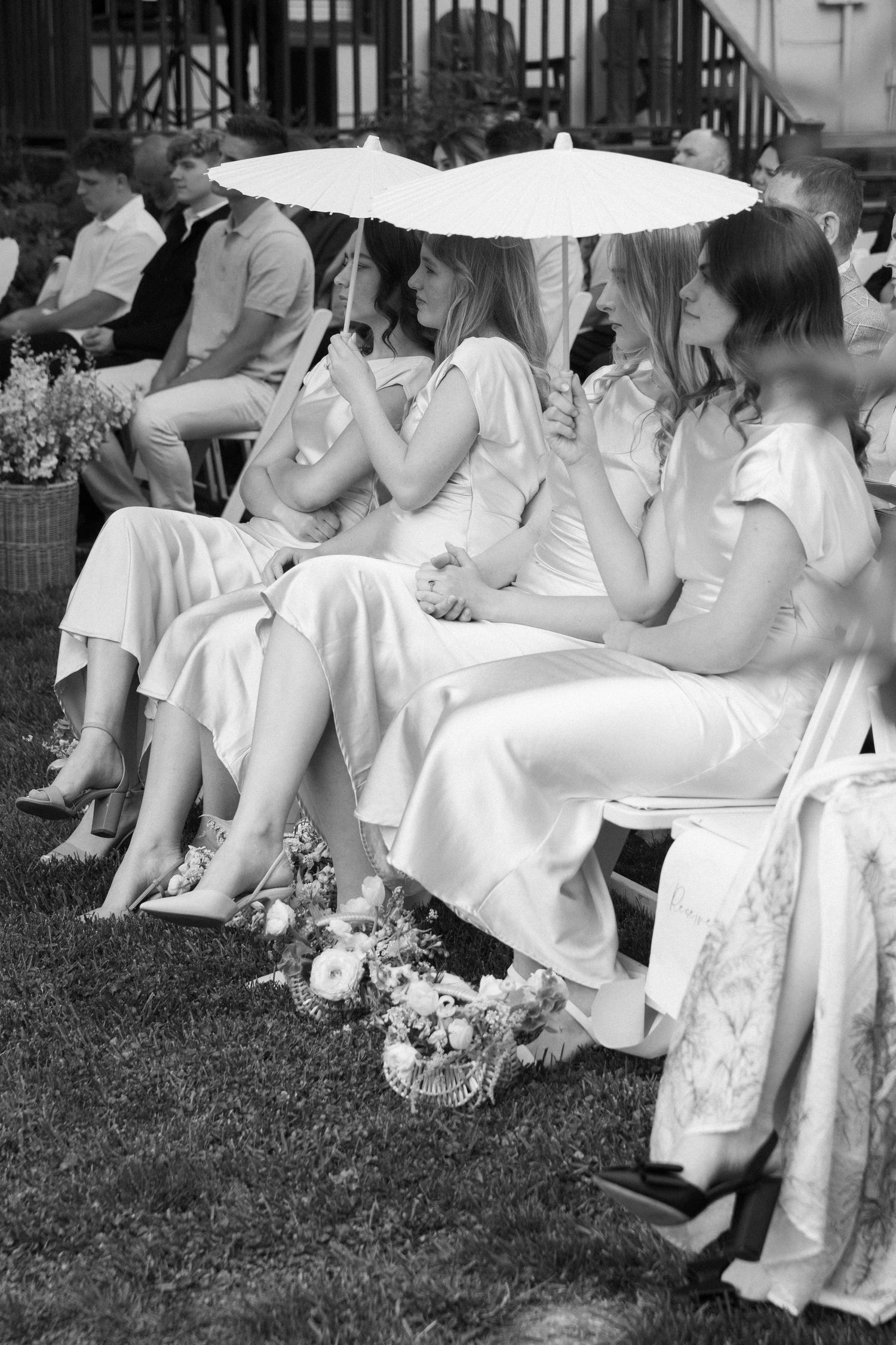 Wedding guests in light dresses holding parasols while seated in white chairs on lawn.