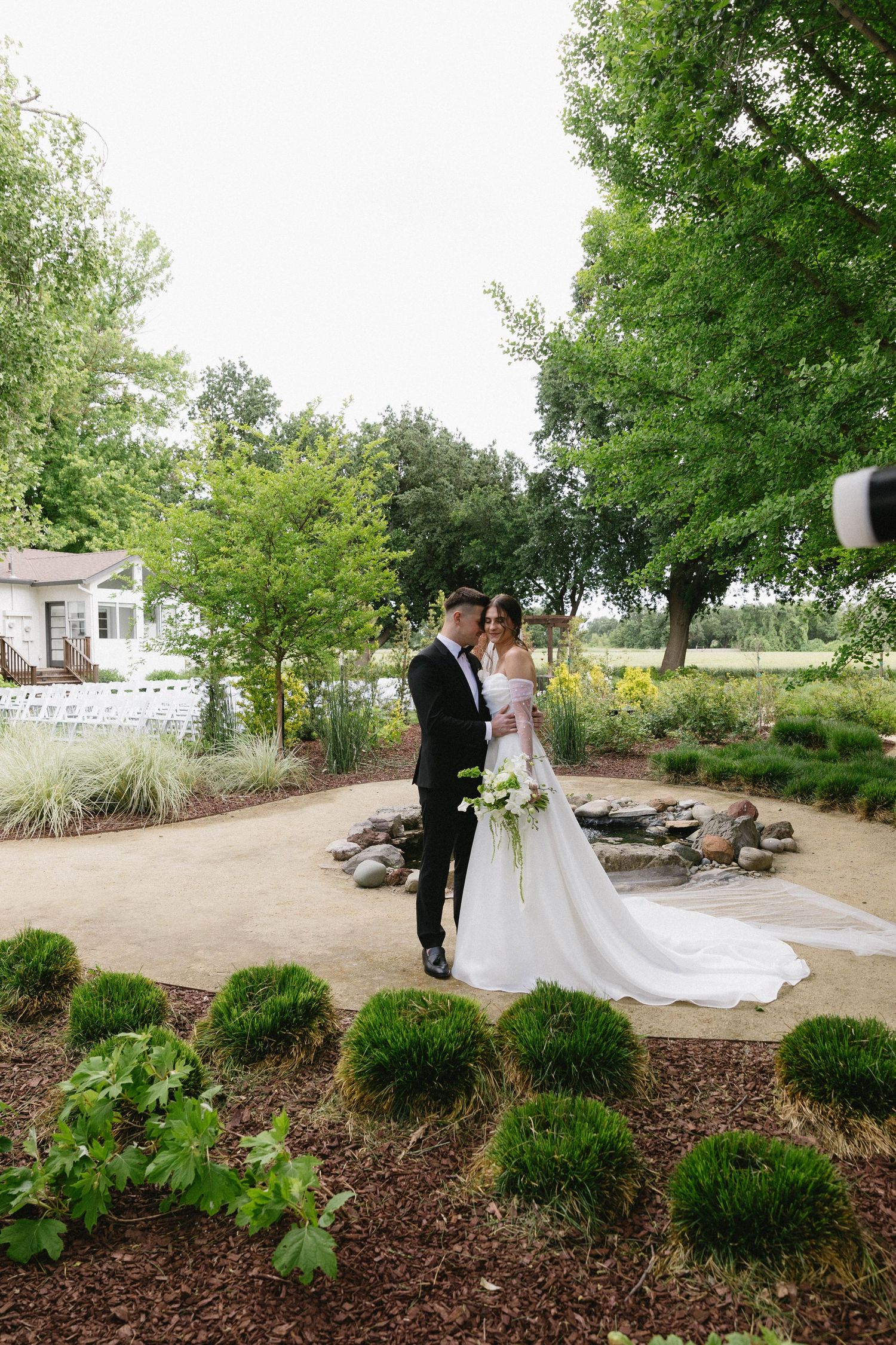A wedding couple poses in a landscaped garden setting with decorative shrubs and mulch.