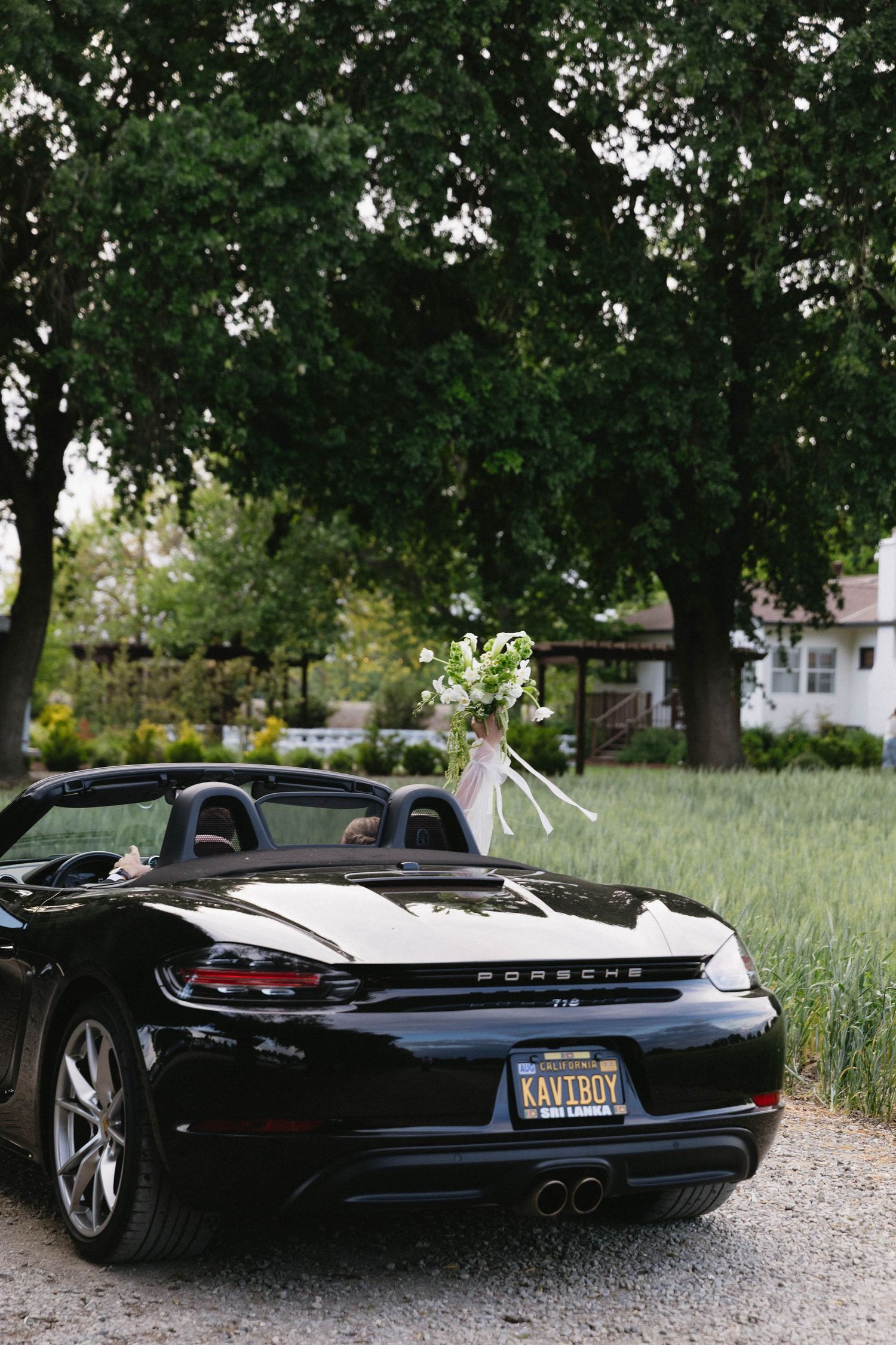 Black Porsche sports car parked under trees on a gravel driveway.